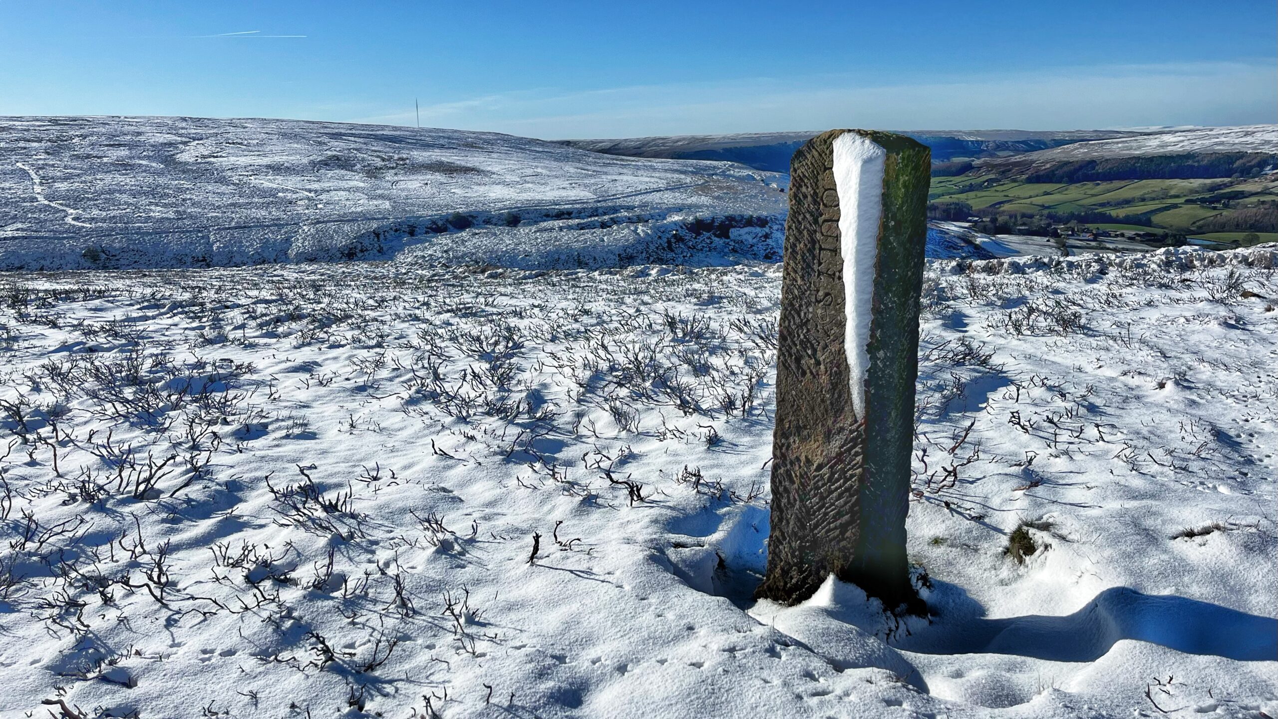 A wide shot of a snowy, moorland landscape under a clear blue sky. In the foreground, a tall, weathered stone boundary marker stands upright. The name "FOULIS" is inscribed vertically down its side, though it is partially obscured by a thick vertical stripe of snow clinging to the stone. The ground is covered in a layer of crisp snow, with dark, low-lying heather and old swiddens poking through. In the background, rolling white hills stretch toward the horizon, and the green valley of Bilsdale is visible in the distance to the right.