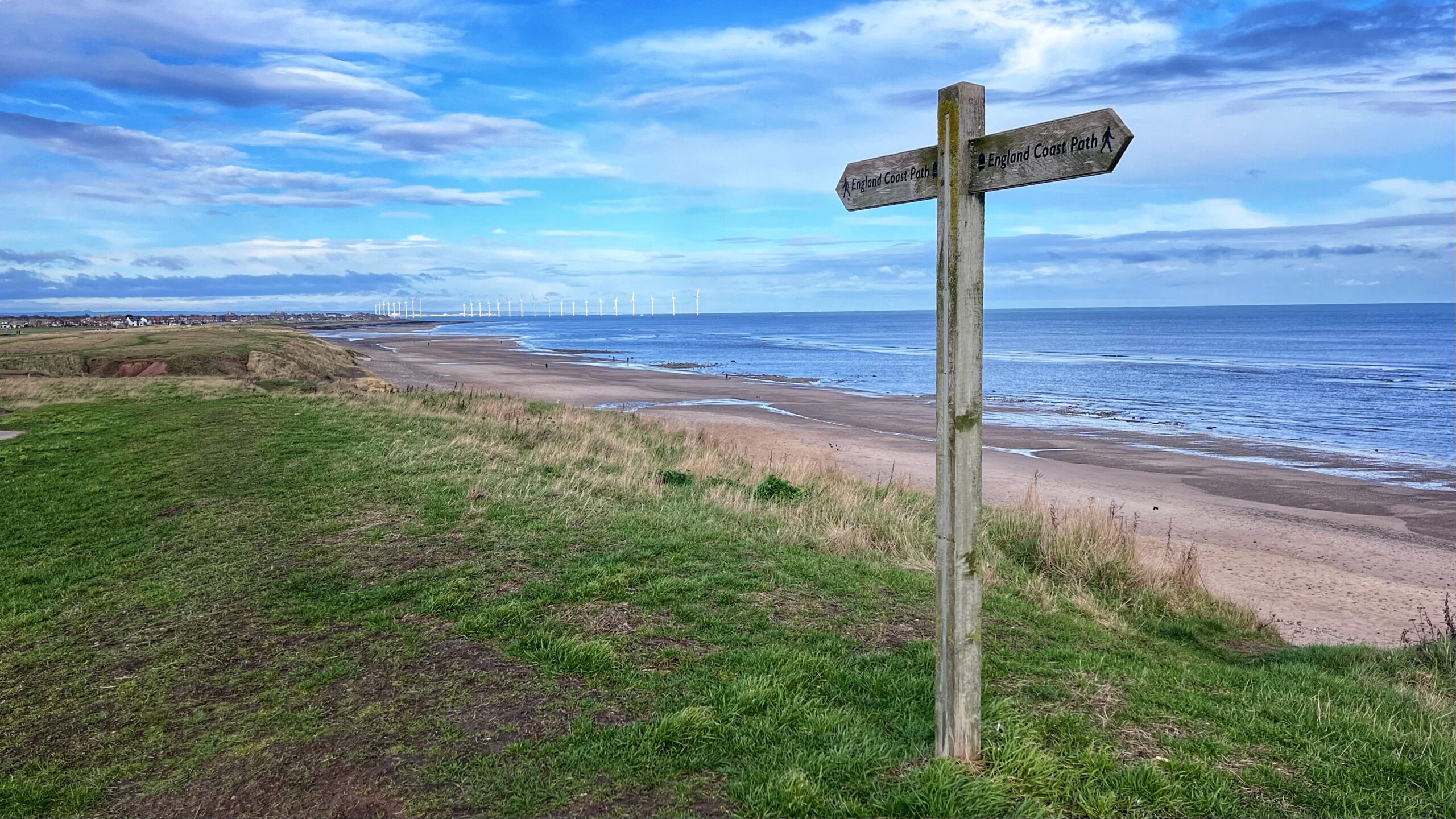 An expansive coastal view from a grassy cliffside overlooking a wide, sandy beach and the North Sea. In the foreground, a wooden fingerpost sign points in two directions along the England Coast Path. The beach curves into the distance toward a town on the horizon, while a long line of white offshore wind turbines stands out against the blue, cloud-streaked sky.