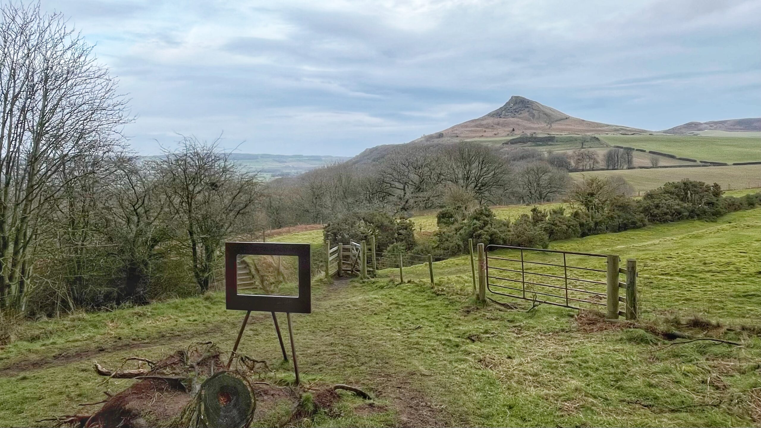A wide landscape view of Roseberry Topping, a prominent conical hill with a jagged cliff edge, under an overcast sky in North Yorkshire. In the foreground, a grassy hillside features a metal gate and a unique rectangular art installation resembling an empty picture frame on a tripod. Bare winter trees and rolling green fields lead toward the hill in the distance.