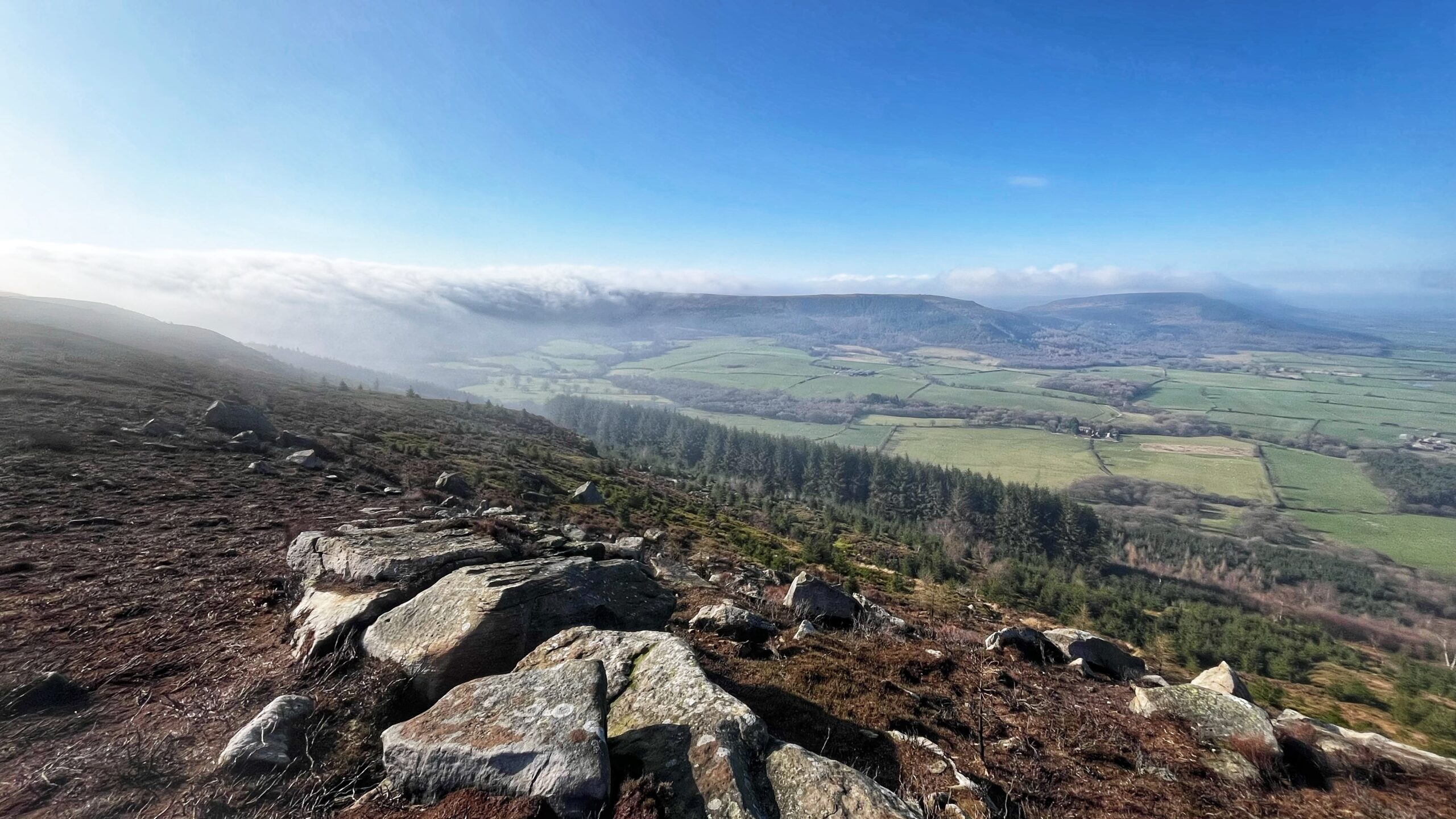 A view from a rocky escarpment on the northern edge of the North York Moors, looking down over Greenhow Bottom. The valley floor is a neat patchwork of green fields divided by hedgerows and dry stone walls, with a belt of dark conifer forest in the middle distance. The flat-topped Cleveland Hills sit opposite under a bright blue sky, with a bank of low cloud rolling over their edge like a white tablecloth sliding off a table. The foreground is burnt heather, peat and scattered sandstone boulders.