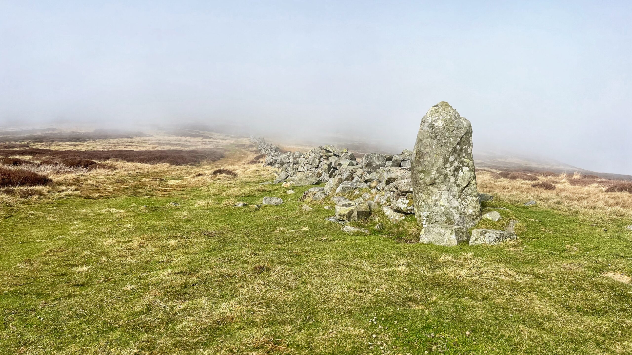 A ruined dry-stone wall stretches across open moorland and disappears into thick hill fog. A large upright standing stone anchors the near end of the wall. The ground is rough moorland grass with patches of dead heather. The sky and horizon are almost entirely swallowed by low cloud.​​​​​​​​​​​​​​​​