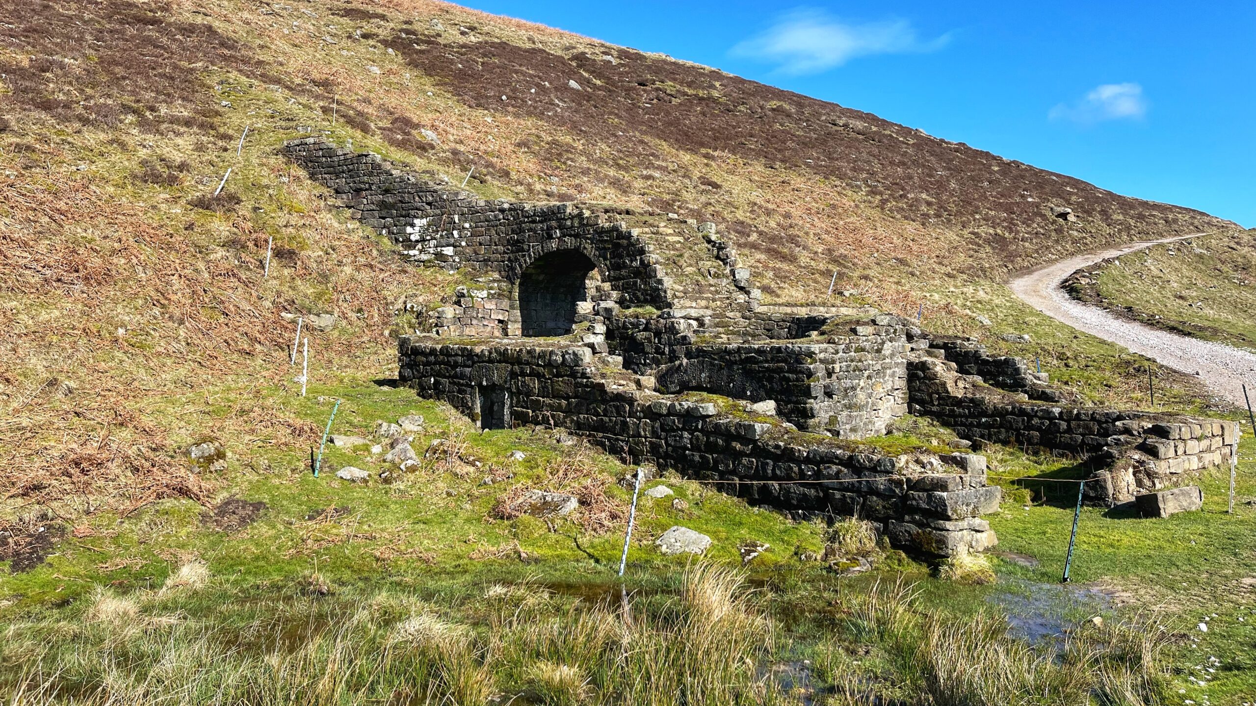 Stone ruins of Providence smelt mill on a moorland hillside, featuring a partially standing arched opening and low crumbling walls of dark dressed stone. Rough grassland and dead bracken surround the site. A gravel track winds up the hill behind it, under a rather optimistic blue sky.​​​​​​​​​​​​​​​​