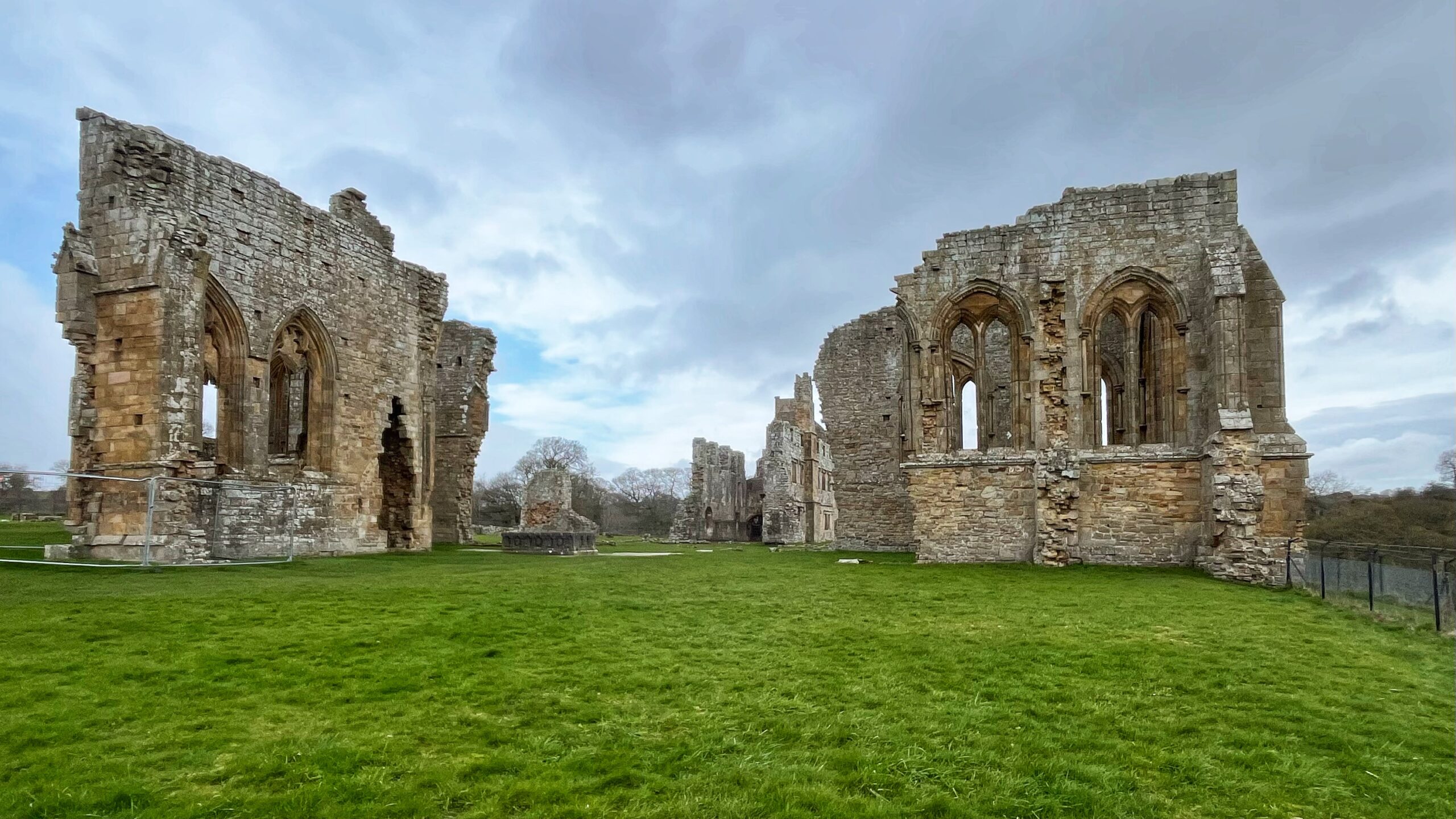 Ruins of Egglestone Abbey, within the historic boundary of the North Riding of Yorkshire. Two tall, roofless stone walls with Gothic arched windows stand on either side of a wide stretch of bright green grass, under a grey, cloudy sky. More crumbling walls and a tower stretch into the distance between them. The stonework is a warm mix of brown and grey. A metal fence is visible on the right.​​​​​​​​​​​​​​​​