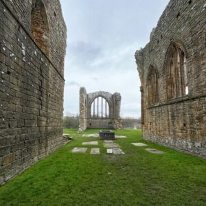 Interior view of the roofless nave of Egglestone Abbey, within the historic boundary of the North Riding of Yorkshire. Tall stone walls rise on both sides, leading the eye towards the remains of a large Gothic east window at the far end, its stone tracery still standing against a pale, overcast sky. Flat grave slabs are set into the bright green grass floor, with a carved stone tomb chest positioned in the centre. The warm brown stonework is patched with lichen and age.​​​​​​​​​​​​​​​​