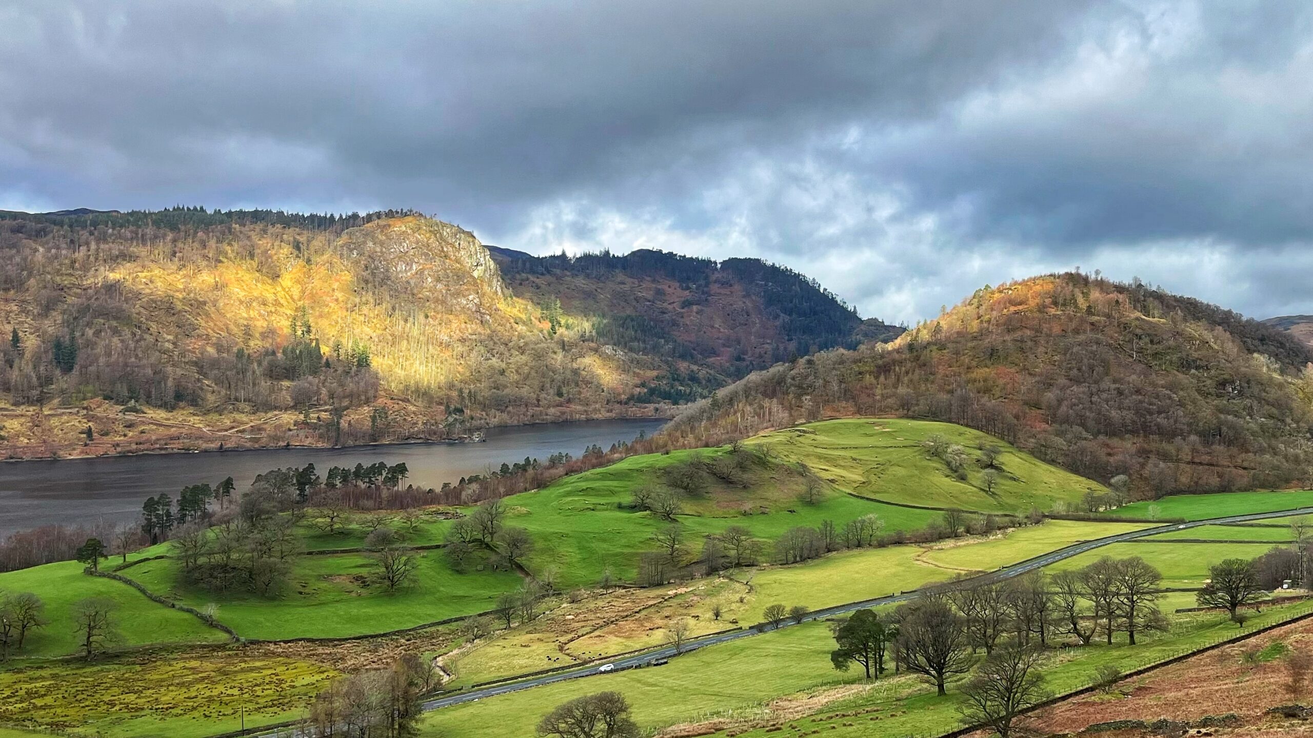 A dramatic Lake District landscape showing Raven Crag dramatically lit by a shaft of golden sunlight against a brooding, storm-grey sky, overlooking the dark, still waters of Thirlmere Reservoir. Rolling green fields divided by dry-stone walls sweep across the valley floor, dotted with bare winter trees. The busy Keswick to Ambleside road powers noisily through the scene, as though nobody told it how spectacular the view was.​​​​​​​​​​​​​​​​