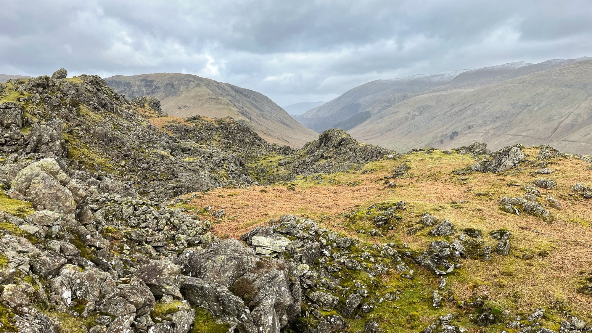 A view across the rocky summit ridge of Helm Crag in the Lake District, showing two parallel lines of dark, lichen-covered boulders and broken rock separated by a hollow of rough tawny grass. Beyond the ridge, a broad glacial valley sweeps away between bare, rounded fells under a heavy grey sky, with a distant hint of snow on the higher ground to the right.​​​​​​​​​​​​​​​​