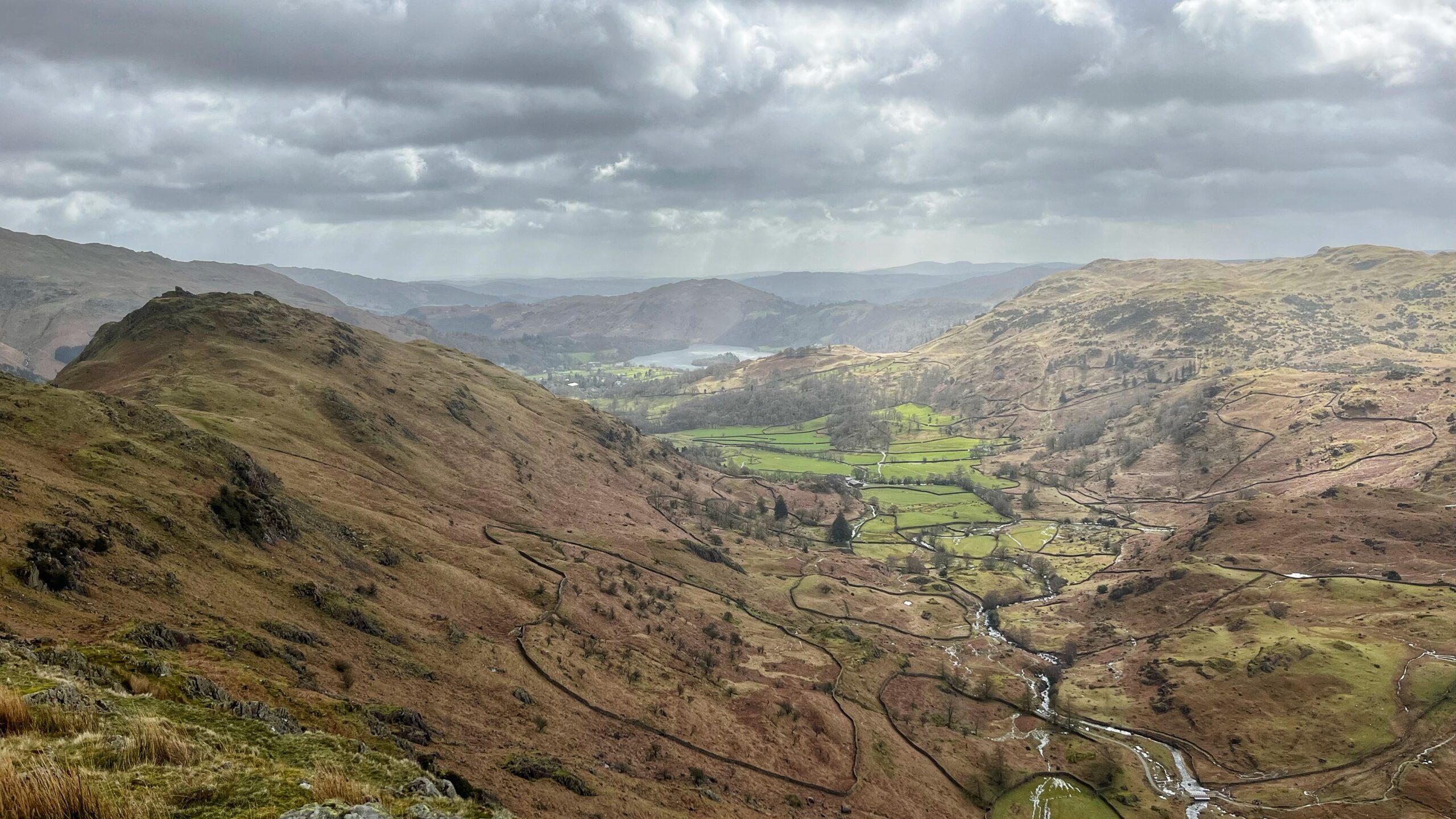 A wide view of Easedale from high on the fells, looking north towards Grasmere lake shimmering faintly in the distance under a brooding grey sky. The valley floor below is a patchwork of bright green fields divided by dark dry-stone walls and many winding tributaries of Easedale Beck. Brown and russet moorland slopes rise on both sides, with the rocky ridge of Helm Crag prominent on the left. Shafts of light break through the clouds over the far hills. Altogether not a bad sort of day for the Lake District.​​​​​​​​​​​​​​​​
