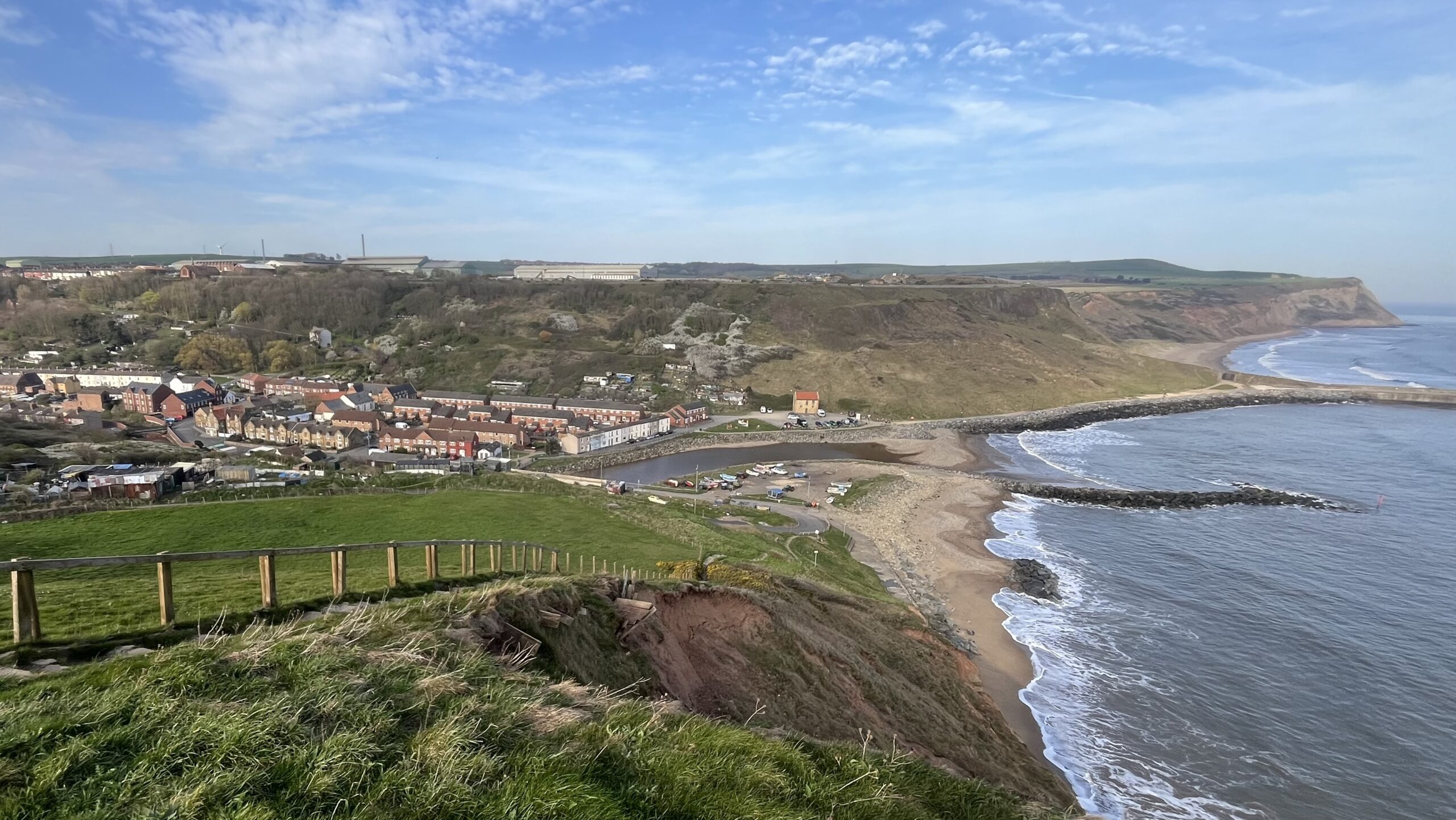 A view from a clifftop above Skinningrove, a small former mining village on the North Yorkshire coast. Terraced houses cluster around a narrow beck that meets a sandy beach. Rocky breakwaters hold back the North Sea, which is doing its level best to reclaim the shore. Rust-red cliffs curve away southward under a pale blue sky. Industrial buildings sit on the horizon, reminding you that this place has always earned its living the hard way. A wooden fence in the foreground is losing an argument with coastal erosion.​​​​​​​​​​​​​​​​