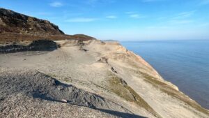 A vast, barren coastal headland stretches toward the sea under a clear blue sky. The ground is pale grey and almost entirely without vegetation, scarred by centuries of industrial alum working. Dark shale and loose rock debris cover the foreground, while the exposed cliff edge drops sharply to the North Sea. A distant headland is visible along the coastline to the right.​​​​​​​​​​​​​​​​