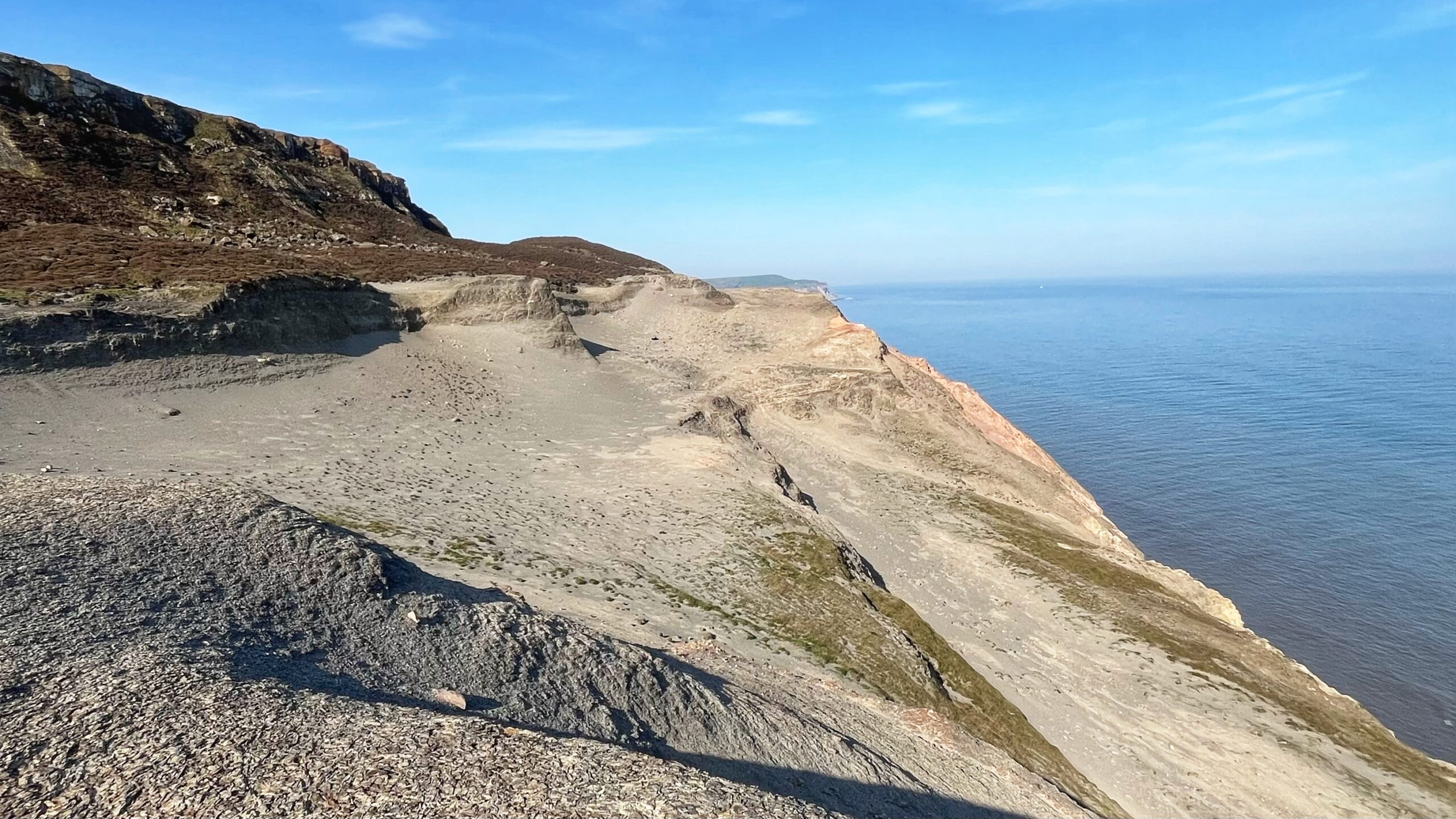 A vast, barren coastal headland stretches toward the sea under a clear blue sky. The ground is pale grey and almost entirely without vegetation, scarred by centuries of industrial alum working. Dark shale and loose rock debris cover the foreground, while the exposed cliff edge drops sharply to the North Sea. A distant headland is visible along the coastline to the right.​​​​​​​​​​​​​​​​