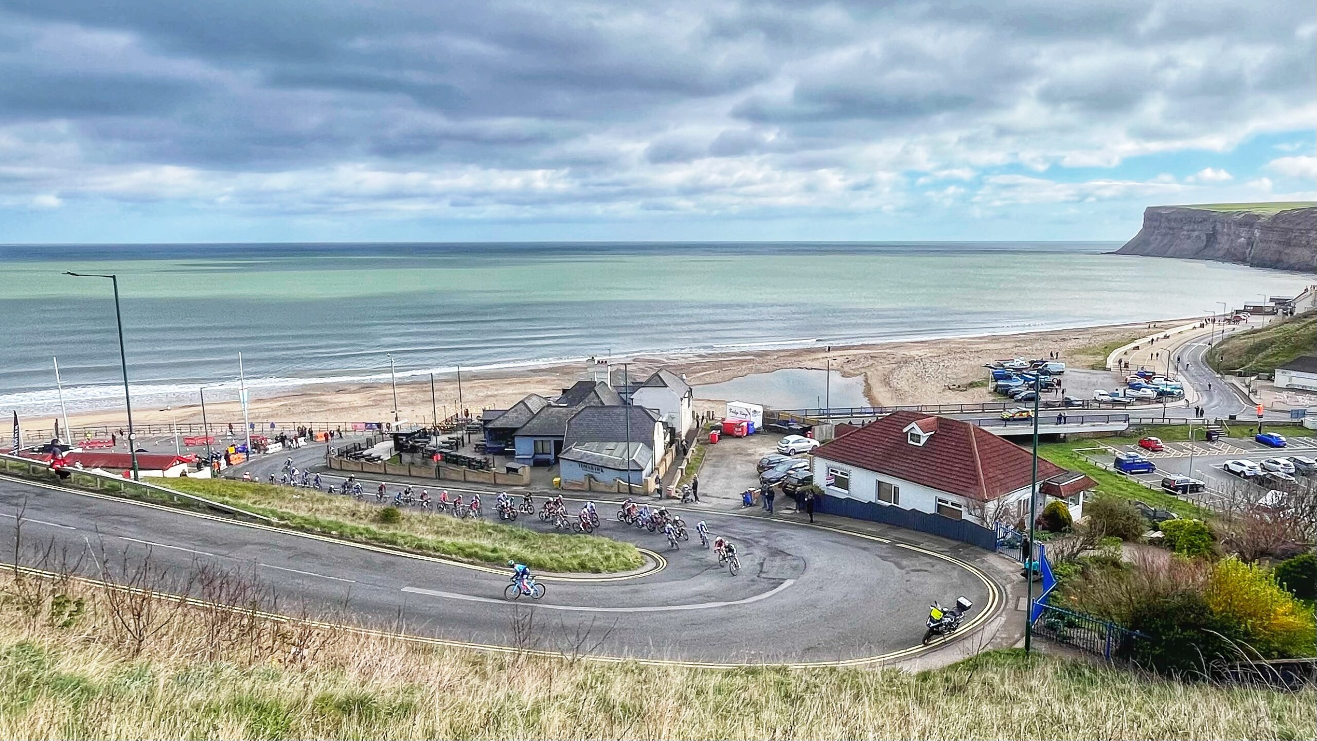A peloton of women cyclists rounds a sharp hairpin bend at the foot of Saltburn Bank, beginning the steep climb away from the seafront. The North Sea stretches flat and grey beneath a heavy, cloud-filled sky, with Saltburn’s sandy beach and dramatic cliffs visible in the background. Small groups of spectators line the roadside near the beachfront buildings as the riders, strung out through the bend, begin their ascent.​​​​​​​​​​​​​​​​