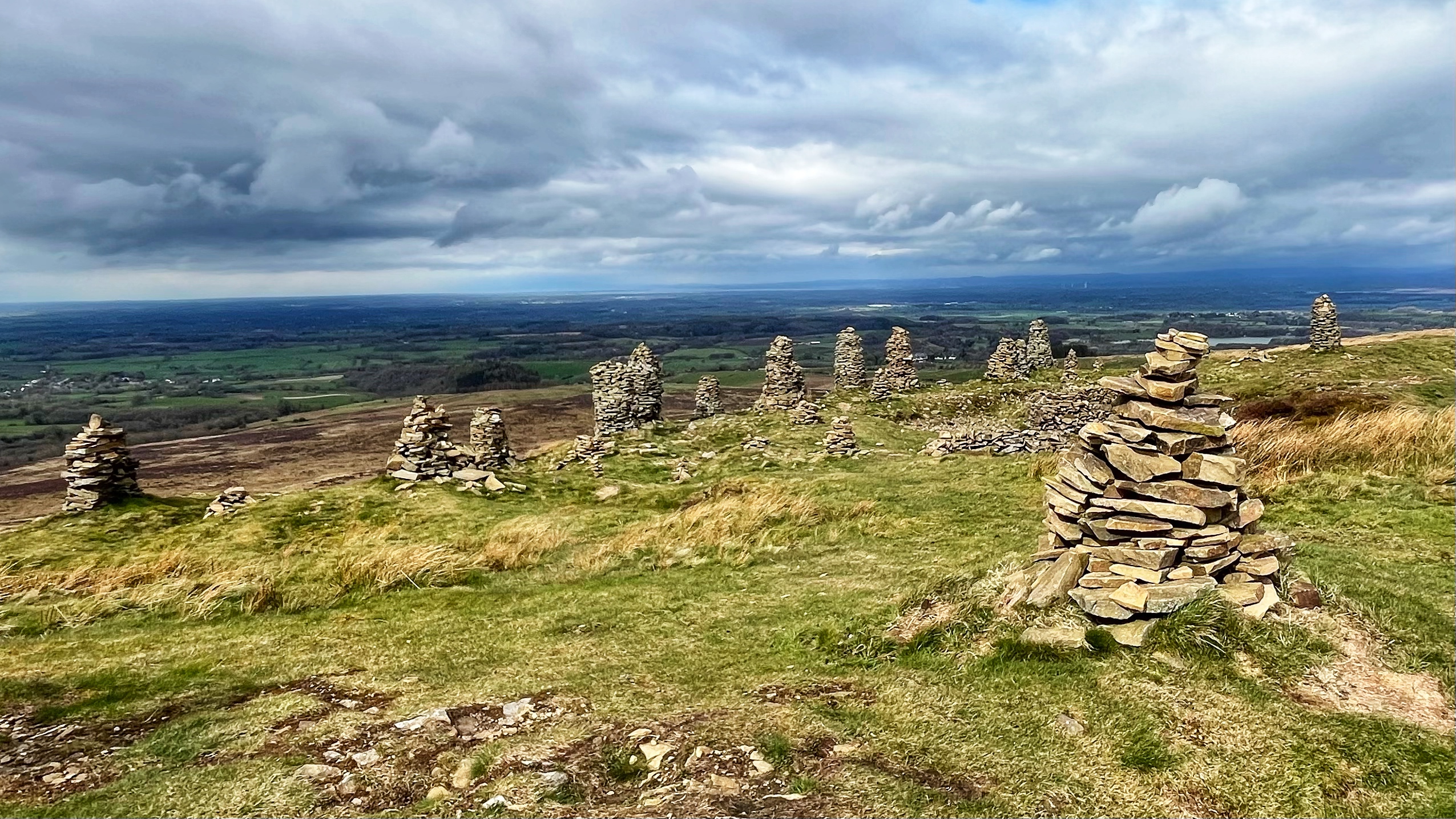 A broad fell summit in Cumbria under a dramatic, cloud-filled sky, with a sweeping panoramic view over a patchwork of green fields, woodland and the distant water of the Solway Firth stretching to the horizon. Scattered across the grassy, tawny hillside in the foreground and middle distance stand more than a dozen dry-stone curricks — tall, neatly stacked pillars of flat sandstone slabs, ranging from roughly half a metre to well over a metre in height. The curricks are spread irregularly across the slope, giving the scene a distinctly ceremonial feel. The nearest currick, in the bottom right of the frame, is not the largest but is the most clearly detailed.​​​​​​​​​​​​​​​​