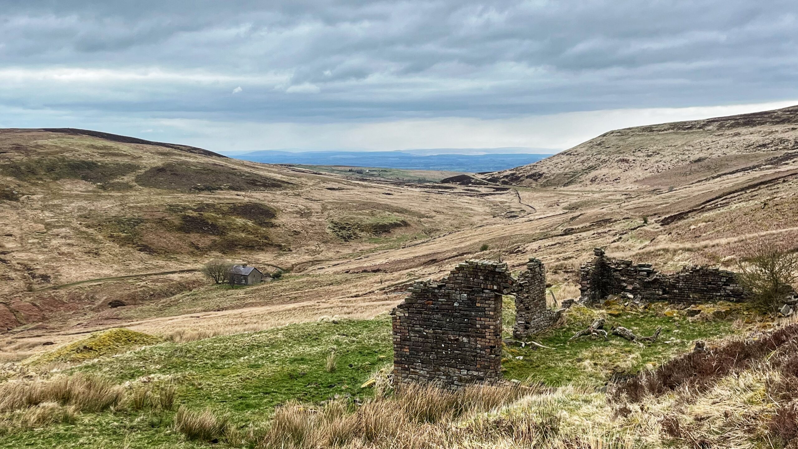 A desolate upland valley in Geltsdale, Cumbria, under a heavy grey sky with patches of pale blue. In the foreground, the roofless brick shell of a ruined building stands on a grassy hillside — the remains of Gairs colliery — its walls crumbling and stained with age and lichen. The valley floor stretches away into the middle distance, covered in rough moorland grasses in shades of brown and ochre, with a winding track visible on the right-hand slope. A lone intact stone building with a grey roof sits further down the valley, sheltered by a handful of bare trees. Beyond the valley mouth, the land opens out to a vast lowland plain, with the faint blue outlines of distant mountains — the North Pennine fells — visible on the horizon.​​​​​​​​​​​​​​​​