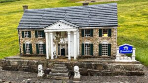 A miniature stone-built replica of Graceland, Elvis Presley’s famous Memphis mansion, sitting in a Cumbrian hillside garden at Nenthead. The building faithfully reproduces the original’s white classical portico with four columns, green window shutters, and front steps flanked by two small stone lions. A blue roadside sign to the right reads “Graceland” in white lettering. The white portico paint is visibly peeling, lending the whole thing a rather melancholy air. Seated casually between the columns is a tiny figure of Elvis himself. Behind and above the model, the unmistakably soggy green hills of the North Pennines roll away into the distance, which is arguably the least likely backdrop Graceland has ever had.​​​​​​​​​​​​​​​​