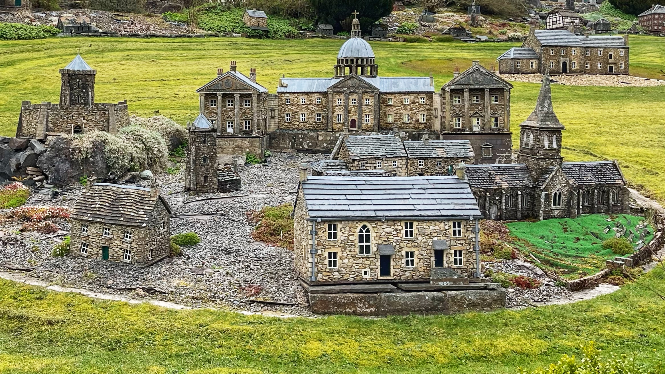 A photograph of a section of Lowson Robinson’s miniature stone village at Nenthead, set on a sloping grass lawn. In the foreground sit two small cottages with slate roofs and tiny white-framed windows. Behind them stands a grand domed building resembling a stately home or civic hall, flanked by a castellated tower to the left and a church with a spire to the right. Further back, a large multi-windowed manor house sits on higher ground. All the buildings are constructed from local stone and built to remarkably fine detail, including miniature chimneys, steps, doorways and slate roofing. The whole scene is rather extraordinary for something sitting quietly in a Cumbrian garden.​​​​​​​​​​​​​​​​