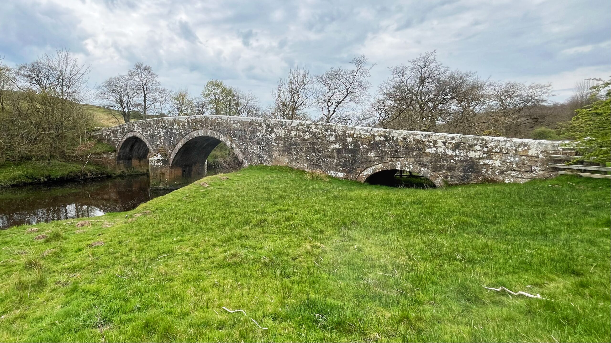 A stone arch bridge built in 1715 spans the River Rede in rural Northumberland. Viewed from the north/west bank, the bridge shows two principal arches over the river and a smaller flood arch on the right bank, all built from rough-cut sandstone heavily encrusted with grey and white lichen. The roadway is low-parapet and almost flat across the top. The river below is calm and dark. The surrounding landscape is vivid spring green grass on both banks, with bare-branched deciduous trees behind the bridge and a hill rising to the left. The sky is bright with broken cloud.​​​​​​​​​​​​​​​​