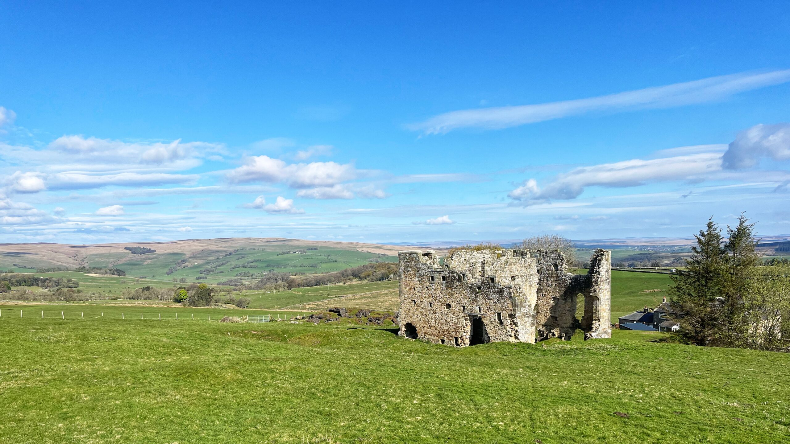 The ruined stone engine house of Ridsdale Ironworks stands on a gently rising green hill under a broad, bright blue Northumberland sky scattered with thin white clouds. The two-storey sandstone shell, open to the sky and missing much of its east wall, shows arched window openings, square machinery holes, and rough rubble edges where Armstrong’s men tore it apart. Rolling green fields and moorland hills stretch away to the horizon behind it, and a cluster of grey-roofed farm buildings sits quietly to the lower right. A single conifer stands to the far right. It looks every inch like a ruined medieval castle, which is rather the point.​​​​​​​​​​​​​​​​