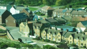 A close aerial view of the centre of Skinningrove village, showing a mix of honey-coloured Yorkshire stone terraces and later brick houses. At the heart of the image stands a notably grand stone building with a cross-shaped footprint, which appears to be a former Co-operative store or similar Victorian village institution — the sort of building that once thought rather a lot of itself. A lone cyclist makes their way along the quiet road below, apparently unbothered by the weight of local history overhead. The flat-roofed Skinningrove Village Hall is visible to the right, suggesting this little village has always looked after its own. Green wooded hillsides press in from all sides.​​​​​​​​​​​​​​​​