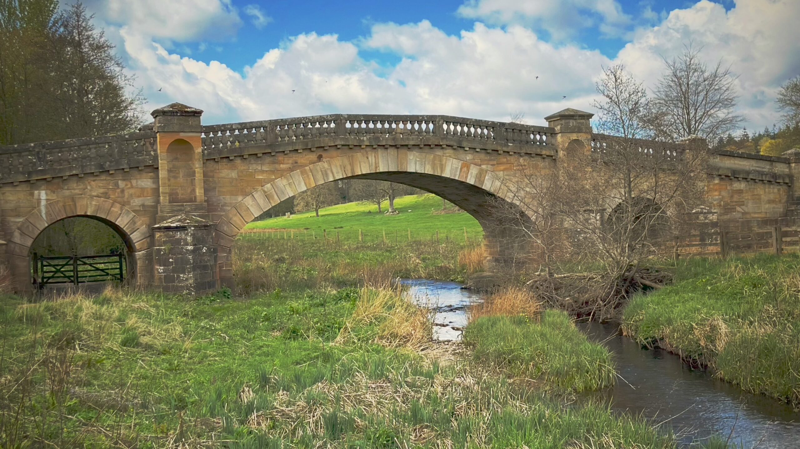 A historic three-arched road bridge built of light-colored, tooled ashlar stone spanning the narrow River Wansbeck in a rural landscape. The bridge features a prominent, high central arch over the water, flanked by two smaller arches over the grassy banks. A decorative stone balustrade runs along the top of the bridge, punctuated by square stone pillars. The bridge is set against a backdrop of rolling green fields, scattered trees, and a blue sky with white clouds.
