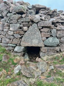 Close view of the broch wall at Culswick, built from large rough pink granite blocks. A small low doorway sits beneath a striking triangular stone lintel, leading into a dark passage within the ruined structure.