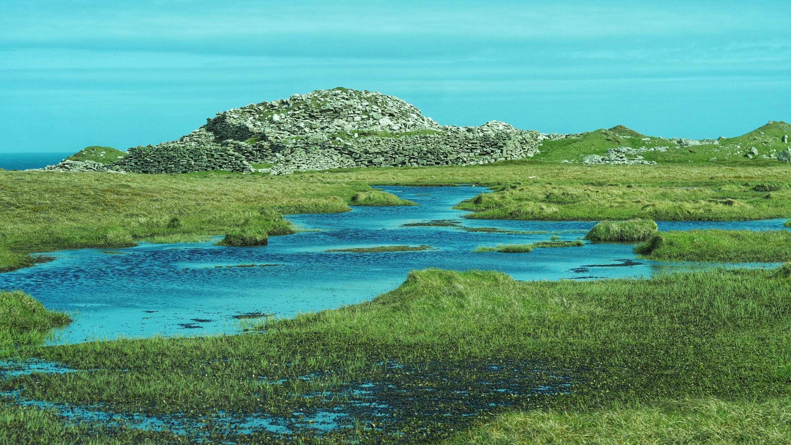A ruined Iron Age broch tower, reduced to a large mound of dry-stone rubble, stands on a green, boggy headland in Levenwick, Shetland. Shallow pools of vivid blue water spread across the waterlogged grassland in the foreground. The sea is just visible to the left, under a bright turquoise sky.