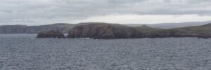 Distant view of the broch at Culswick from the headland at Westerwick, with dark cliffs and rocky headlands stretching beneath a heavy grey sky. Even in its ruinous state, the ancient site dominates the coastline.