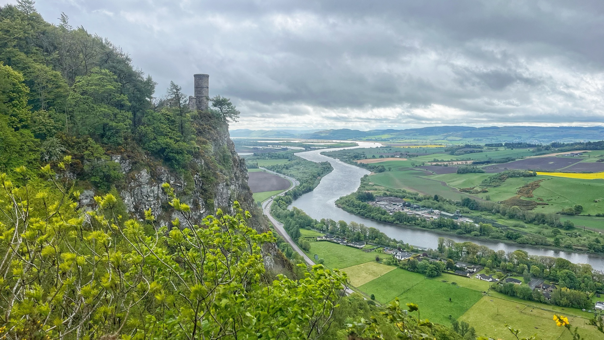 A ruined stone tower perches on the edge of a sheer cliff above the winding River Tay, surrounded by lush green woodland and spring foliage. Rolling farmland, patches of yellow rapeseed, and a winding road stretch across the valley below, all under a suitably moody Scottish sky.