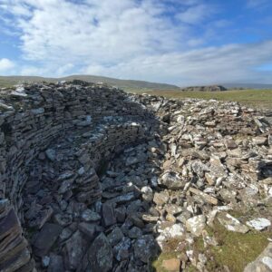 A close-up view looking down into the partially collapsed interior of the Levenwick broch, Shetland. The curved dry-stone wall, built from dark layered flagstone and speckled with grey and white lichen, still stands to a reasonable height on the left. To the right, the wall has collapsed into a jumble of fallen stones. Beyond the ruin, a wide open moorland landscape stretches to distant hills and dramatic coastal cliffs under a blue sky with rolling clouds.