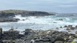 A wild, rocky Atlantic coastline on an overcast day, with white-capped waves crashing against dark, lichen-covered rocks in the foreground and a low headland beyond. The sea is a rather lively turquoise-grey, and a distant island sits on the horizon beneath a pale, sulking sky.