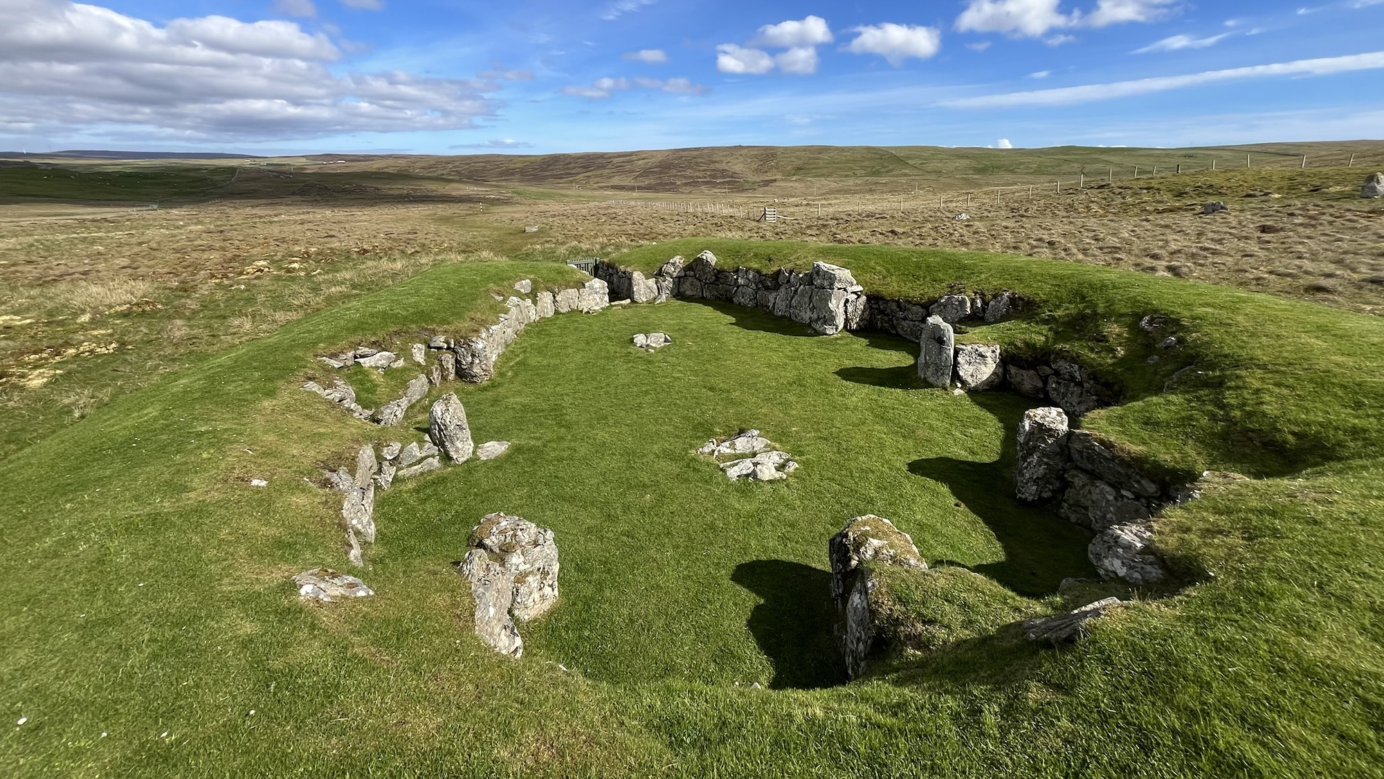A high-angle view of the Neolithic Stanydale Temple in Shetland’s West Mainland, showing its unique heel-shaped plan. The structure features massive walls built from substantial, undressed stone blocks capped with green turf. The interior is a large oval grassy space featuring shallow stone-lined recesses along the rear of the chamber. The temple is set within a vast, treeless moorland landscape under a blue sky with light clouds.