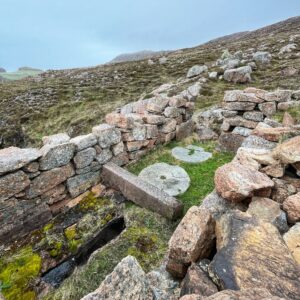 The interior of a roofless, ruinous horizontal watermill (clack mill) constructed from thick pink granite blocks. On the uneven working floor, a pair of original circular millstones (grinding stones) remain, though the wooden fittings and thatched roof have long since decayed. The rugged, dry-stone walls frame a view of the wild, treeless Shetland landscape outside.