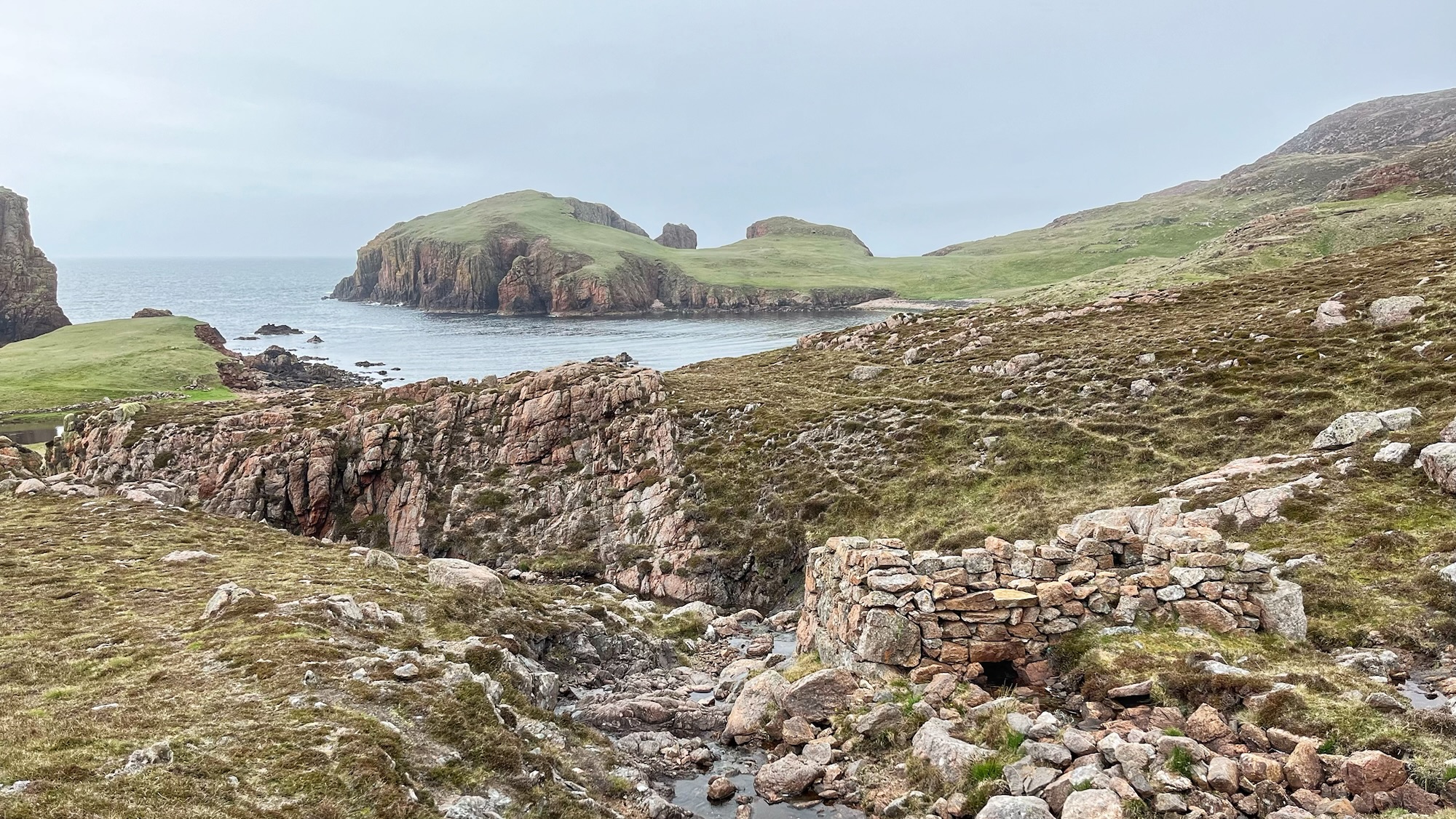 A traditional clack mill (horizontal watermill) featuring rugged dry-stone walls built in pink granite by a narrow, rocky stream. The mill is situated in the wild, treeless landscape of Muckle Roe, an island off Shetland Mainland. In the background, the waters of the sheltered bay of North Ham are visible, framed by the costal cliffs under a wide sky.
