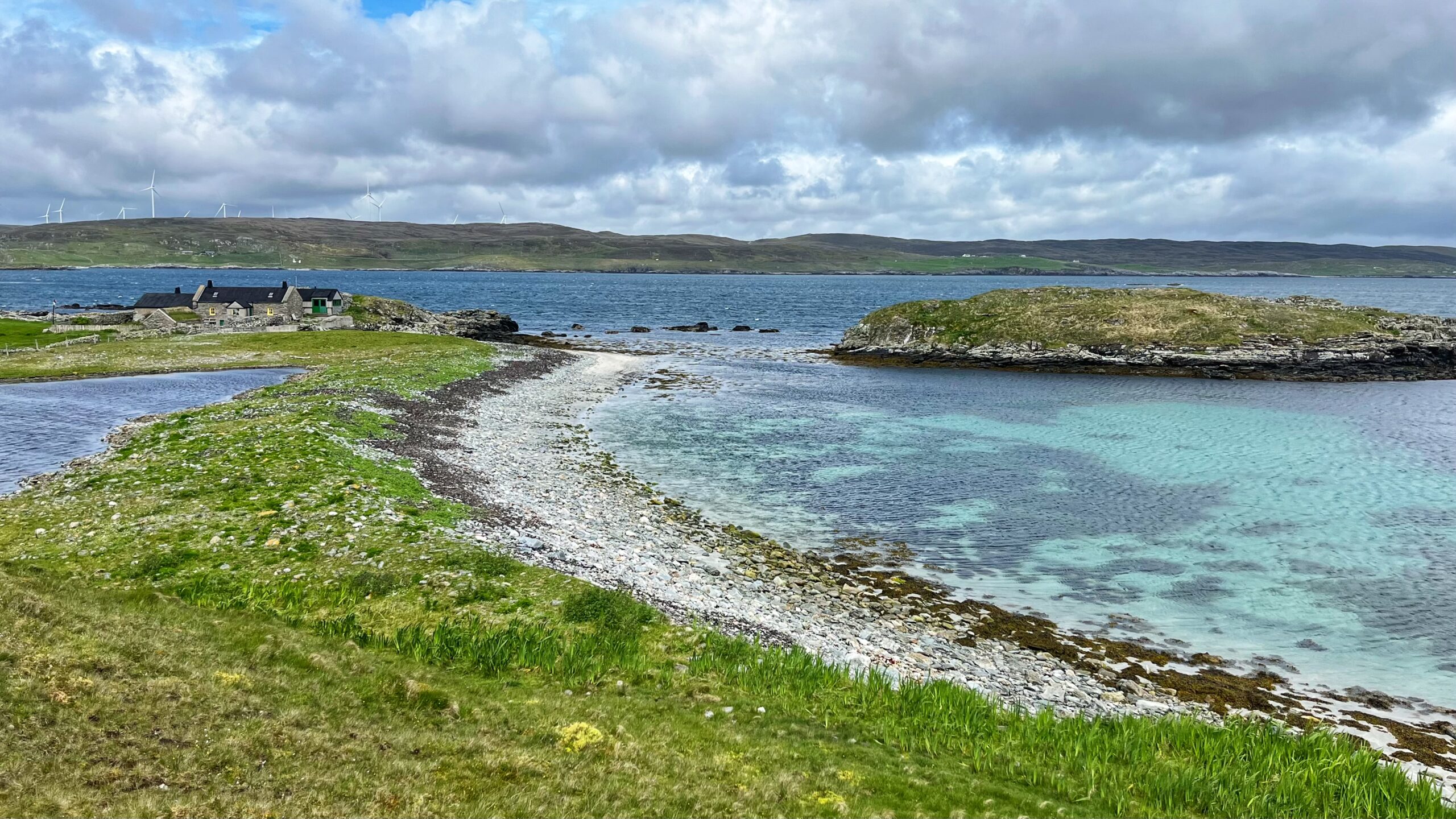 Colour photograph of Ling Ness, Shetland. A curved shingle and shell-sand bar separates a shallow brackish lagoon of clear turquoise water from a broader voe beyond. Short green turf lines the foreground. A small cluster of stone buildings sits to the left, and a rocky islet breaks the water to the right. Wind turbines stand on the low moorland ridge across the water under a wide, cloudy sky.