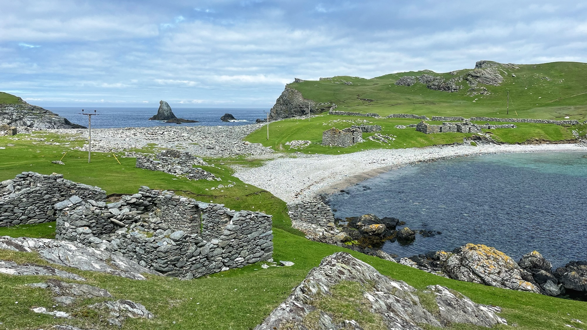 A sweeping view of Fethaland, Shetland, showing the ruins of dry-stone walls and old stone buildings scattered across vivid green grass along a sheltered shingle beach. A calm, dark bay sits to the right, with rocky outcrops and a dramatic sea stack rising from the Atlantic beyond. Rolling green headlands stretch into the distance under a wide, grey-blue sky.