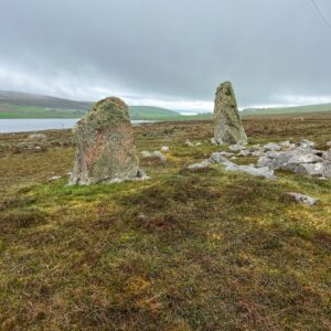 Two ancient standing stones rise from a flat expanse of rough moorland grass and peat, heavily encrusted with lichen and moss, their reddish granite just visible beneath. Between and around them, the scattered remains of a chambered cairn lie low to the ground. A loch stretches across the middle distance, with green rolling hills and a small settlement beyond, all under a brooding, heavy grey sky entirely in keeping with a dreich Shetland morning.