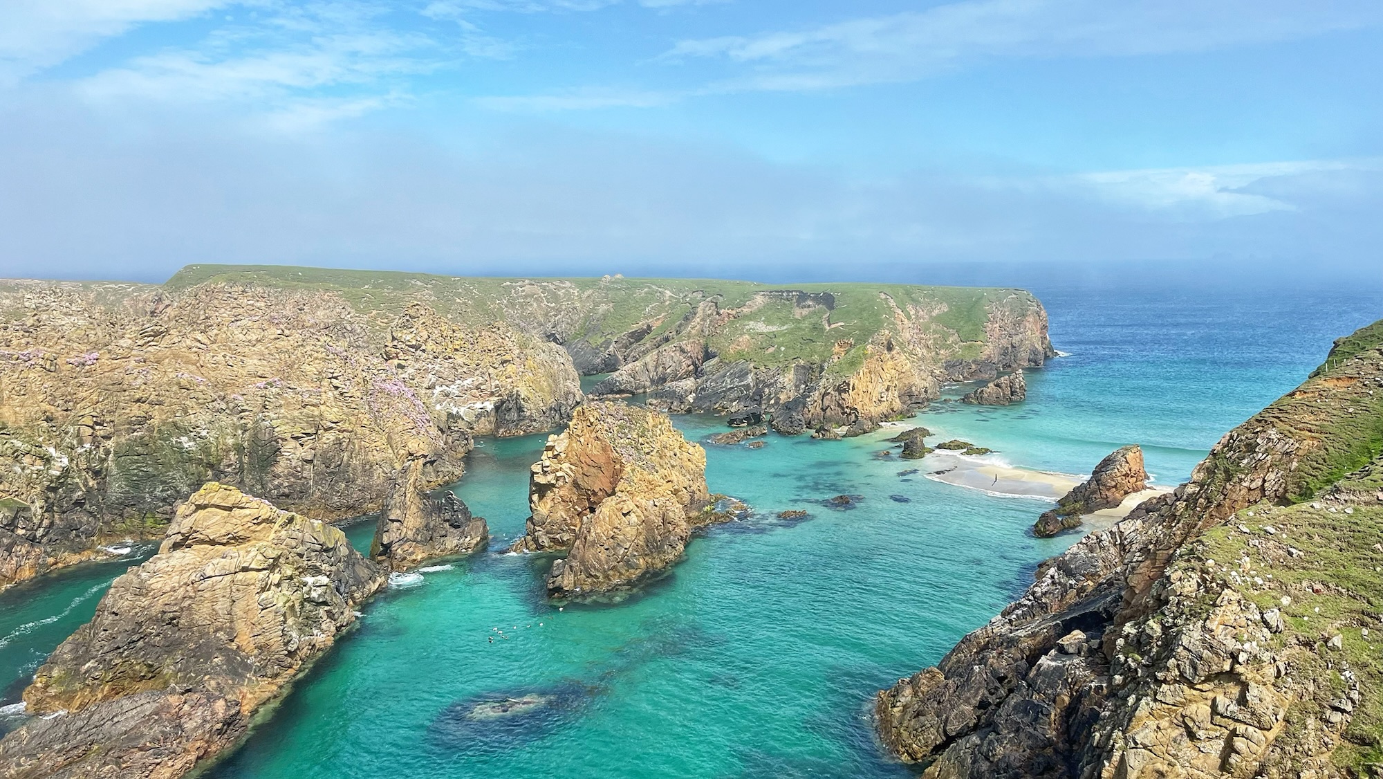 A dramatic coastal scene showing a narrow sound flanked by rugged golden-brown rock stacks and cliff faces dusted with pink sea thrift. The water is a striking turquoise and emerald green, impossibly clear. In the middle distance, a narrow sandy isthmus reaches out towards the island of Uyea, exposed at low tide and offering a brief, tempting crossing point. Green headland stretches across the background under a pale blue sky still carrying traces of morning mist.