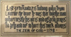 A cream-coloured panel bearing a transcription of the original late 16th-century Scots inscription from above the door of Muness Castle, set in decorative blackletter script within a double-ruled border. The text reads: “List ye to knaw yis bulding quha began / Laurence the bruce he was that worthe man / quha ernestly his airis and ofsprig prayis / To help and not to hurt this derk always / THE ZEIR OF GOD 1598.” The spelling is gloriously unhinged by modern standards, but the meaning is plain enough: please do not wreck my castle.