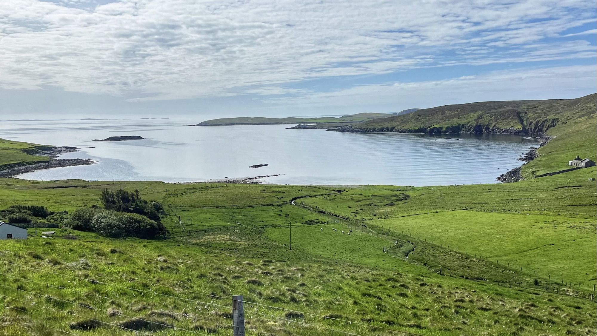 A wide-angle landscape view looking down onto Otterswick (Otters Wick) on the island of Yell, Shetland. In the foreground, a wire fence runs along a vibrant green hillside that slopes down toward a lush coastal valley where a small burn (stream) winds through fields. A few Shetland sheep graze in the pastures below. The middle ground features the calm, blue-grey waters of the sheltered bay (wick), bordered by rolling green headlands and featuring a small dark rock or skerry. On the right-hand shore, a small, solitary grey stone building—characteristic of the island's traditional architecture—sits near the water's edge. The horizon shows distant islands and headlands under a bright sky with soft, hazy horizontal cloud layers.