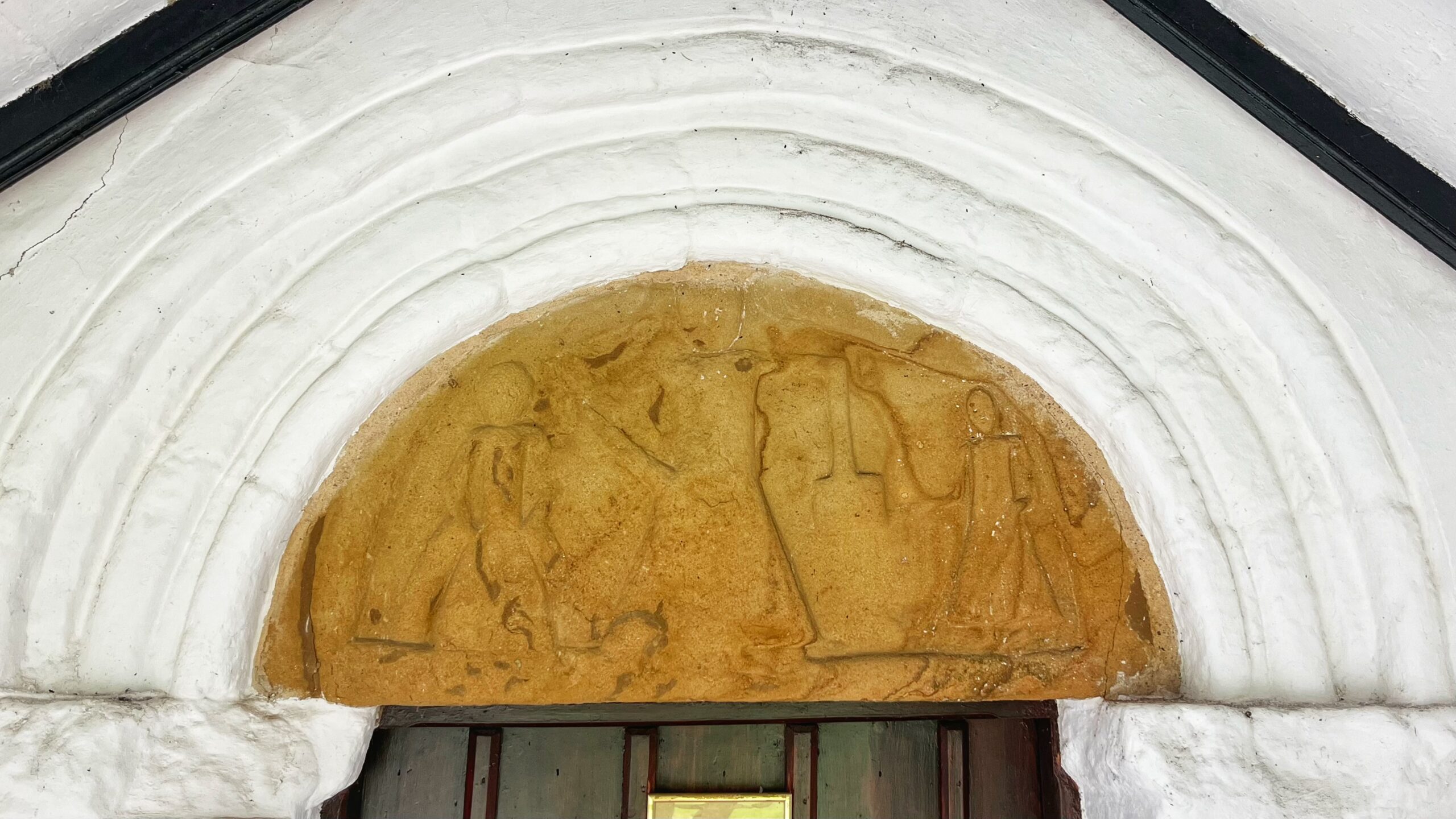 A Norman tympanum of honey-gold sandstone fills the semicircular space above the church door, framed by a whitewashed arch of concentric roll mouldings. The carving is badly worn but is reported to represent three standing men in long robes, one of whom is holding a square object.