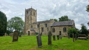 A medieval sandstone church with a square, battlemented tower stands under a heavy grey sky. The nave and side aisle show Gothic pointed windows with leaded glass. A graveyard of tilting and upright stone headstones occupies the foreground, set in close-cropped green grass. Mature trees frame the building on both sides. A modern slate-roofed extension sits to the right of the main body. The stonework is rough-cut and varied in tone, from pale gold to dark brown.