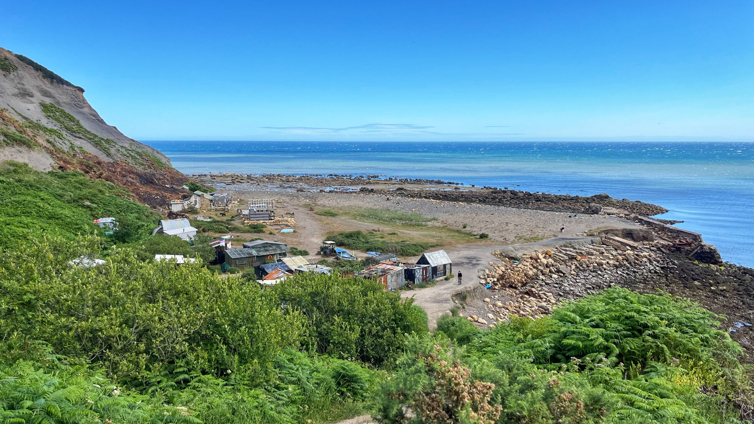A steep grassy and eroded cliff drops down to a pebble beach. At the bottom sits a cluster of weathered wooden and corrugated metal huts, some rusted, with a small boat and an old tractor parked nearby. The tide is out, exposing flat rocky shore. Beyond, the sea stretches to a clear horizon under a blue sky with faint clouds. Two people walk along the shoreline.