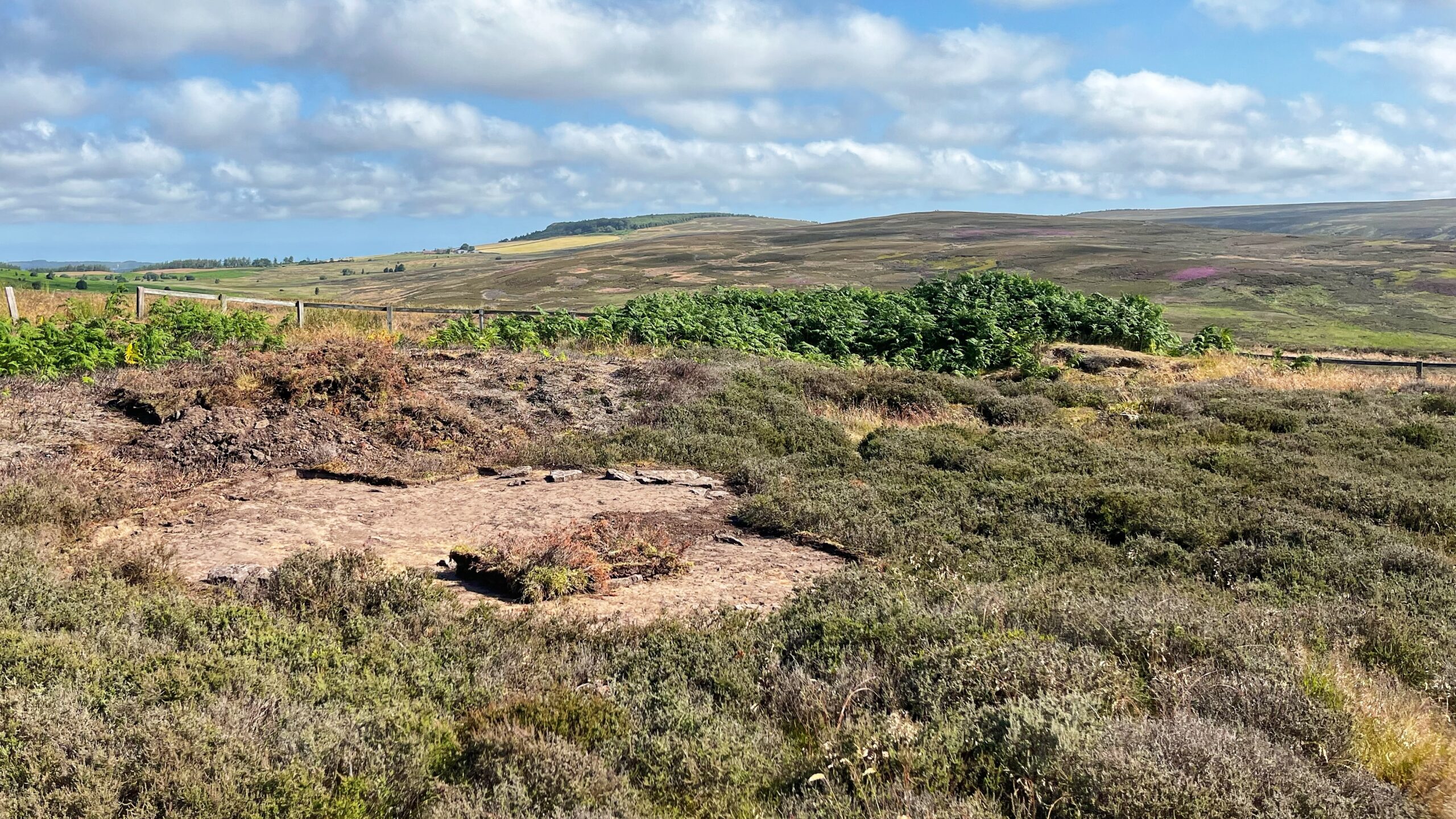 Bare patch of stripped moorland on Percy Rigg, Kildale, showing exposed stone slabs from a hut circle floor among cut heather and bracken. Rolling moorland and distant hills stretch behind under a partly cloudy sky, with a faint glimpse of the coast to the left.