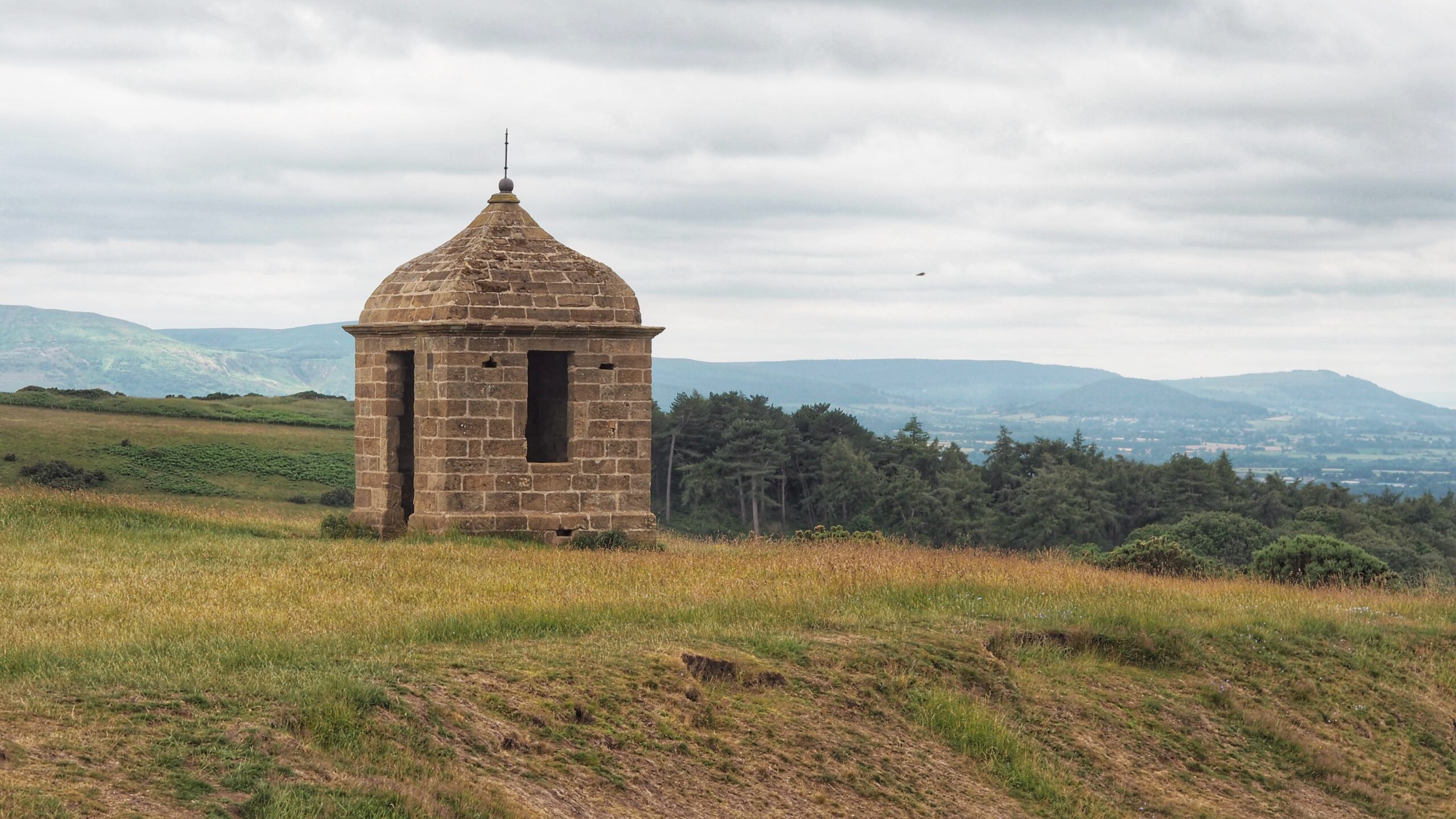 A small stone building with a domed roof and a spike on top, standing alone on a grassy hilltop. The stone has gone the colour of old toffee. Two dark openings serve as doorways, empty and staring like eyes with nothing behind them. Behind it, a slope of pine trees drops away towards a wide valley, with hazy hills rolling off into the distance under a flat grey sky.