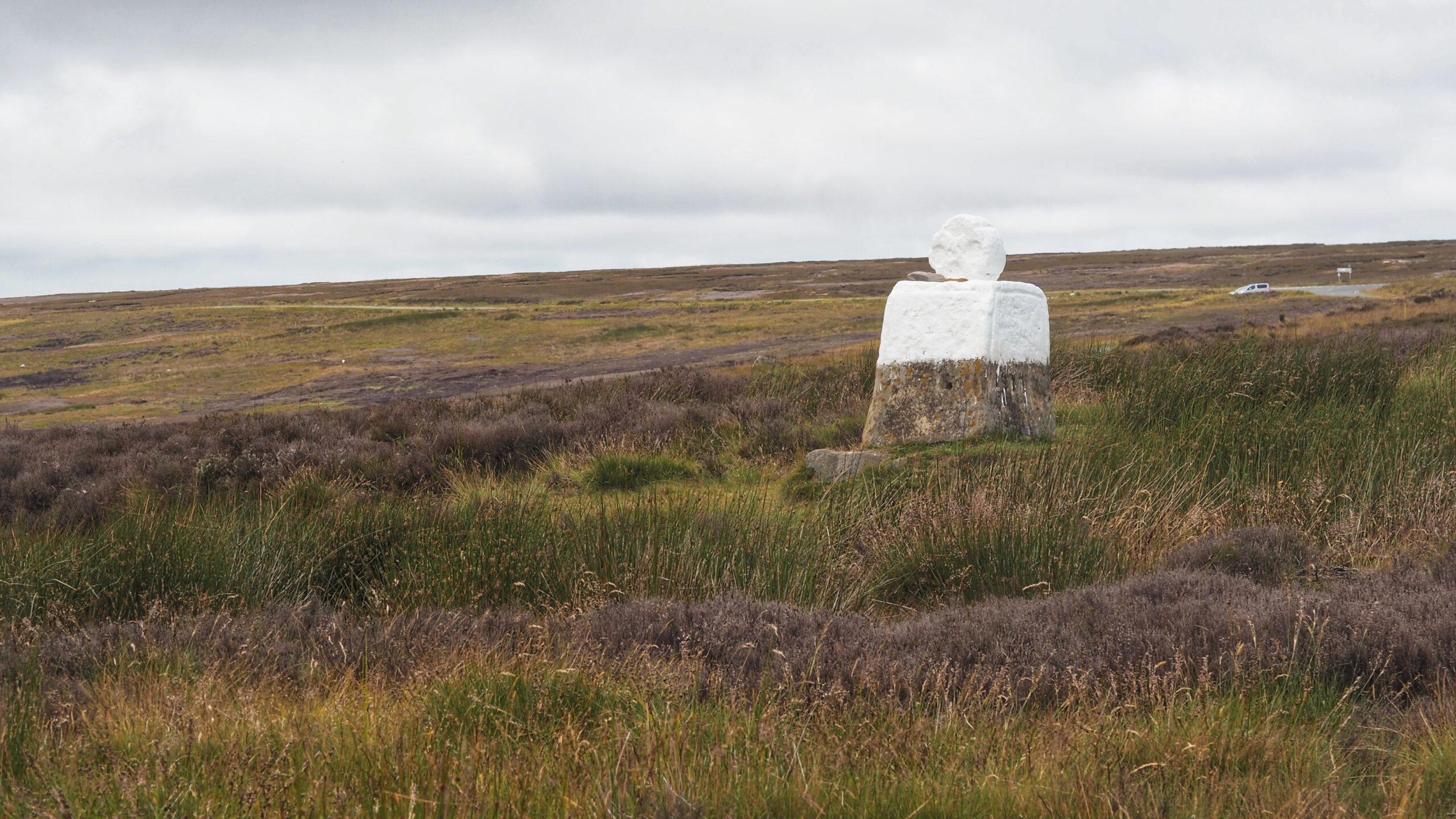 Fat Betty on Danby Moor, seen from a distance across rough heather and grass. A short whitewashed stone base sits below a round white wheelhead, resting slightly off centre on top. The lower part of the base is bare grey stone, weathered and lichen-marked. Open moorland stretches out behind under a flat grey sky, with a narrow road, a parked van, and a small signpost visible in the far distance.