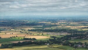 A wide-angle, long-range photograph capturing the southern periphery of Great Ayton village. The subject is a large, light green arable field—the site of the proposed Easby Lane residential development—located just right of the image centre. The field is bounded to the northwest by the established gardens and rooftops of a late 20th-century housing estate and to the southwest by the stretch of Easby Lane. The surrounding environment features a broad agricultural landscape with open fields to the south and east, illustrating the village's historic relationship with its rural setting. In the mid-distance, the wooded banks of the River Leven and the historic core of the village are visible, framed by a vast landscape extending toward the horizon under an overcast sky.