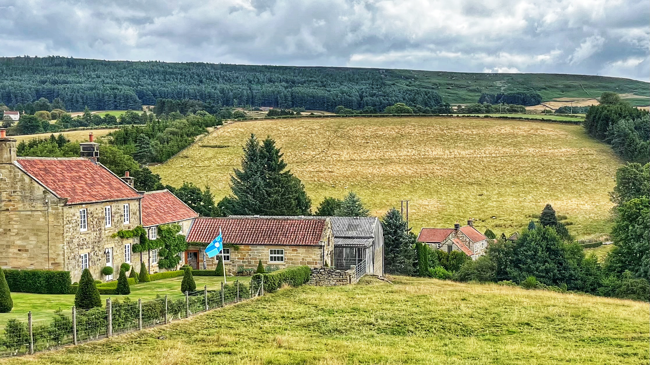A stone farmhouse with red-tiled roofs sits in a valley, the North Riding of Yorkshire flag flying from a pole in the garden. Rolling fields, a dark stretch of woodland, and moorland hills rise beyond under a cloudy sky.