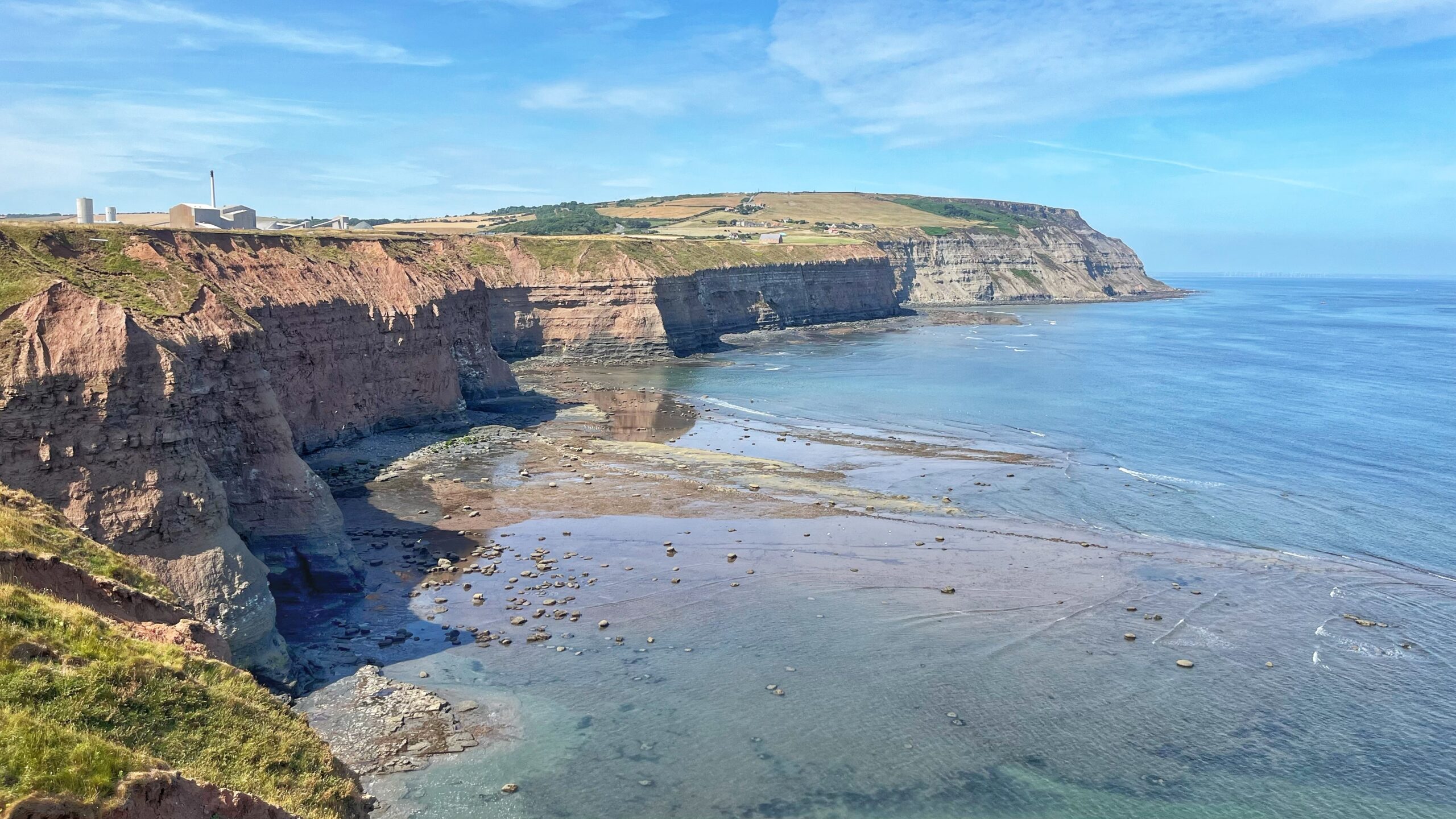 Cliff-top view of the North Yorkshire coast at Hole Wyke, near Staithes. Layered red and grey sandstone cliffs run along the left side of the frame, dropping to a rocky, tide-washed beach exposed at low water. Boulby Mine's buildings and chimney sit on the headland above, with farmland stretching behind. The sea fills the right side under a pale blue sky with light cloud.