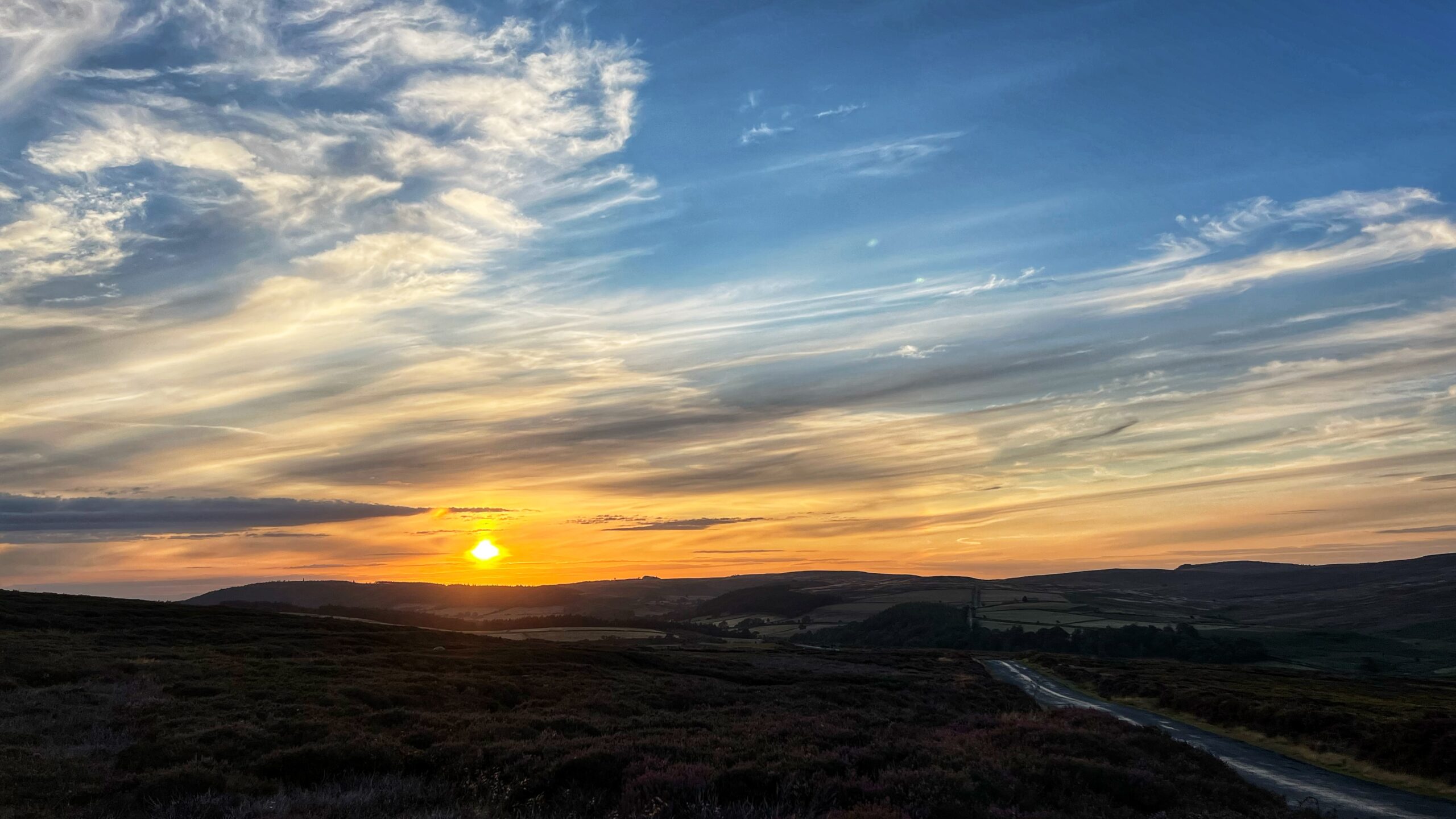 Sunset over rolling moorland hills in the UK, with dark silhouetted heather-covered slopes in the foreground and a bright orange sun low on the horizon. Streaks of cirrus cloud fan out across a blue sky above, catching gold and pink light. A narrow tarmac road curves through the bottom right of the frame, its surface reflecting the last of the evening light.