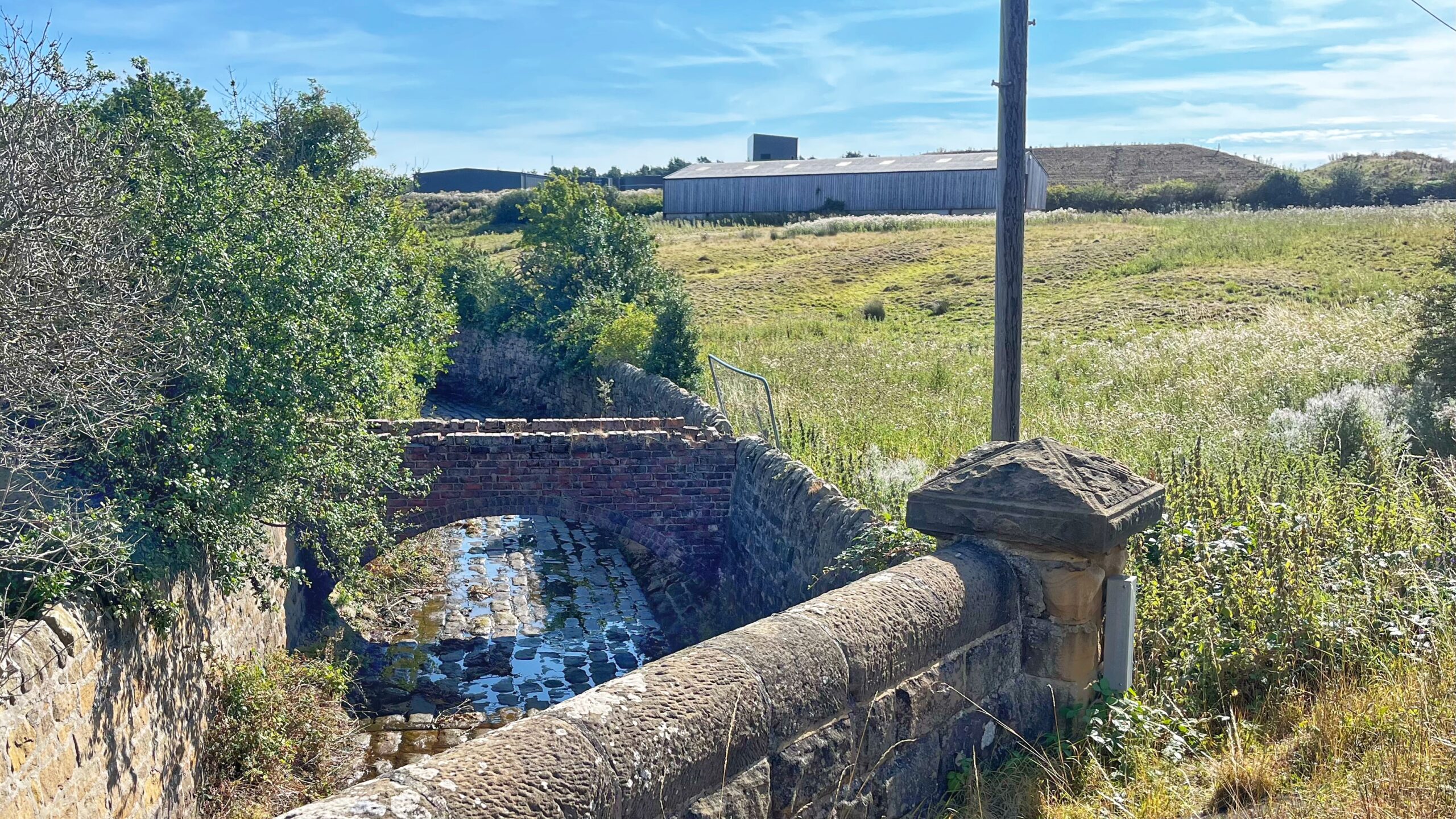 A stone bridge with a brick arch carries a track over a small culvert at Lockwood Beck Reservoir, the stream below running shallow over cobbles. A weathered stone parapet for another bridge stands in the foreground, with rough grassland, a wooden barn and outbuildings visible beyond, under a pale blue sky.