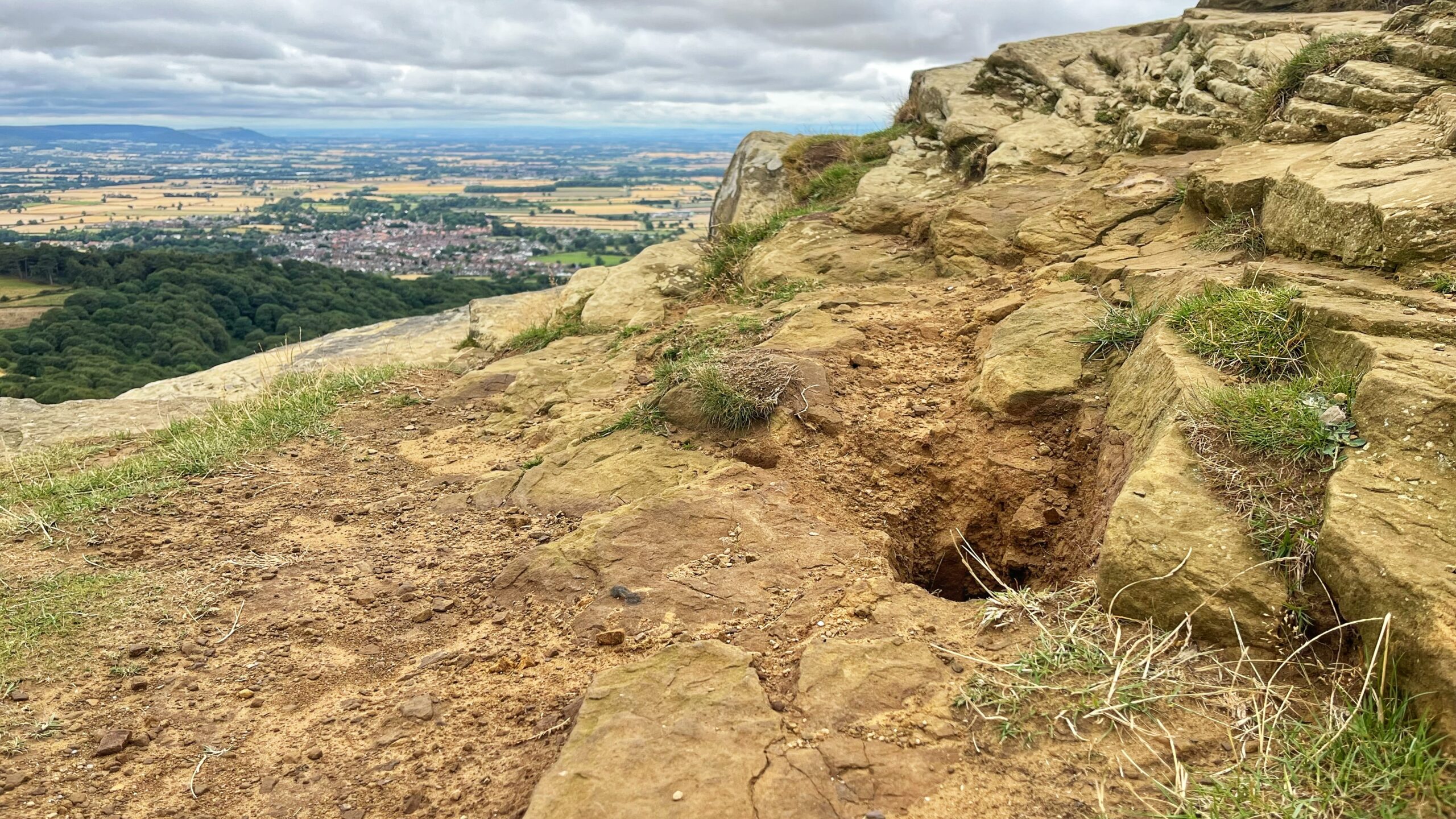Rocky summit of Roseberry Topping with a substantial subsidence hole and eroded ground in the foreground, surrounded by uneven sandstone slabs and tufts of grass. The wooded hillside and patchwork farmland of the valley are visible beyond beneath a cloudy sky.